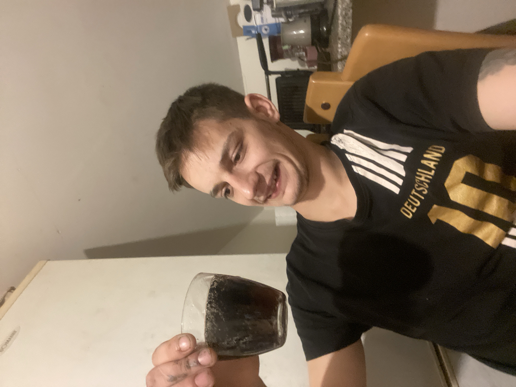 A young man with short brown hair and light skin is holding up a glass of dark liquid with bubbles in his left hand. He is wearing a black t-shirt with white stripes and gold lettering that reads "DEUTSCHLAND 10". His facial expression is one of slight disgust or discomfort, with his mouth slightly open and his eyebrows furrowed. He is seated in a brown chair in what appears to be a domestic setting, with a white refrigerator visible in the background and a countertop with various items behind him. The lighting is dim, suggesting an indoor environment.