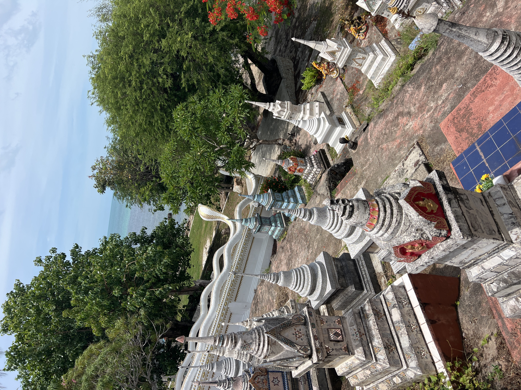 This image depicts a hillside cemetery in Baan Hua Thanon, Thailand. Numerous ornate stupas, varying in size and design, are scattered across a reddish-brown, weathered stone pathway. Some stupas are white and undecorated, while others are adorned with intricate patterns, mosaic tiles, and colorful embellishments. Lush green trees with dense foliage surround the cemetery, and in the distance, the turquoise ocean is visible under a bright, partially cloudy sky. A white, serpent-like railing with a dragon's head snakes along a raised platform in the background, adding to the temple-like atmosphere. Red flowers bloom on a tree in the right foreground.