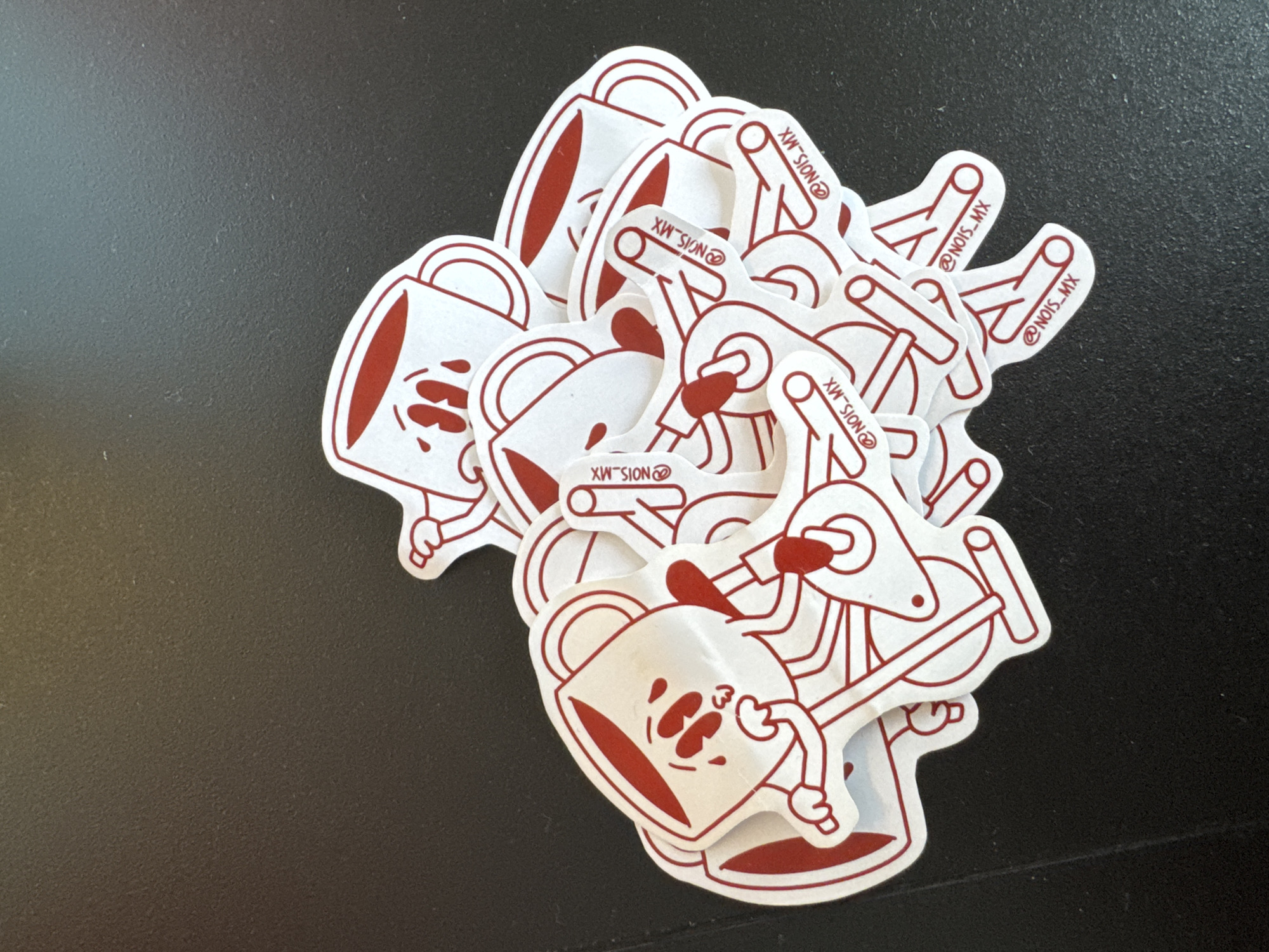 A pile of white stickers with red line art sits on a black surface. The stickers depict two distinct designs: one is a cartoonish coffee mug with a face, and the other is a stylized image resembling a hand-pumped liquid dispenser or gas pump, with the text "@NOIS_MX" incorporated into the design. No people, interactions, or specific environmental details beyond the black surface are visible. The image does not contain sufficient information to identify the location of San Bernardino Tlaxcalancingo, Mexico.