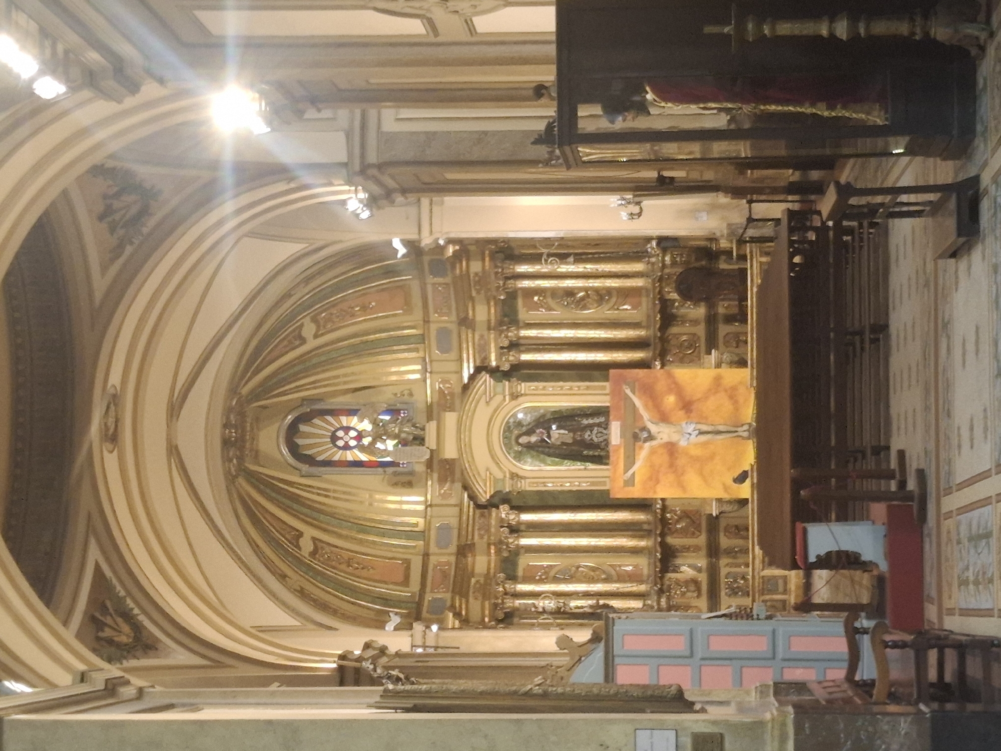 The image shows the interior of a church, likely in Buenos Aires, Argentina. The architecture is ornate, featuring a gilded altar and a curved ceiling with decorative elements. An image of Christ on the cross is prominent to the right of the altar. Wooden organ pipes are visible on the right side of the image. A small table and chair are also visible, suggesting an area for a choir or musicians.