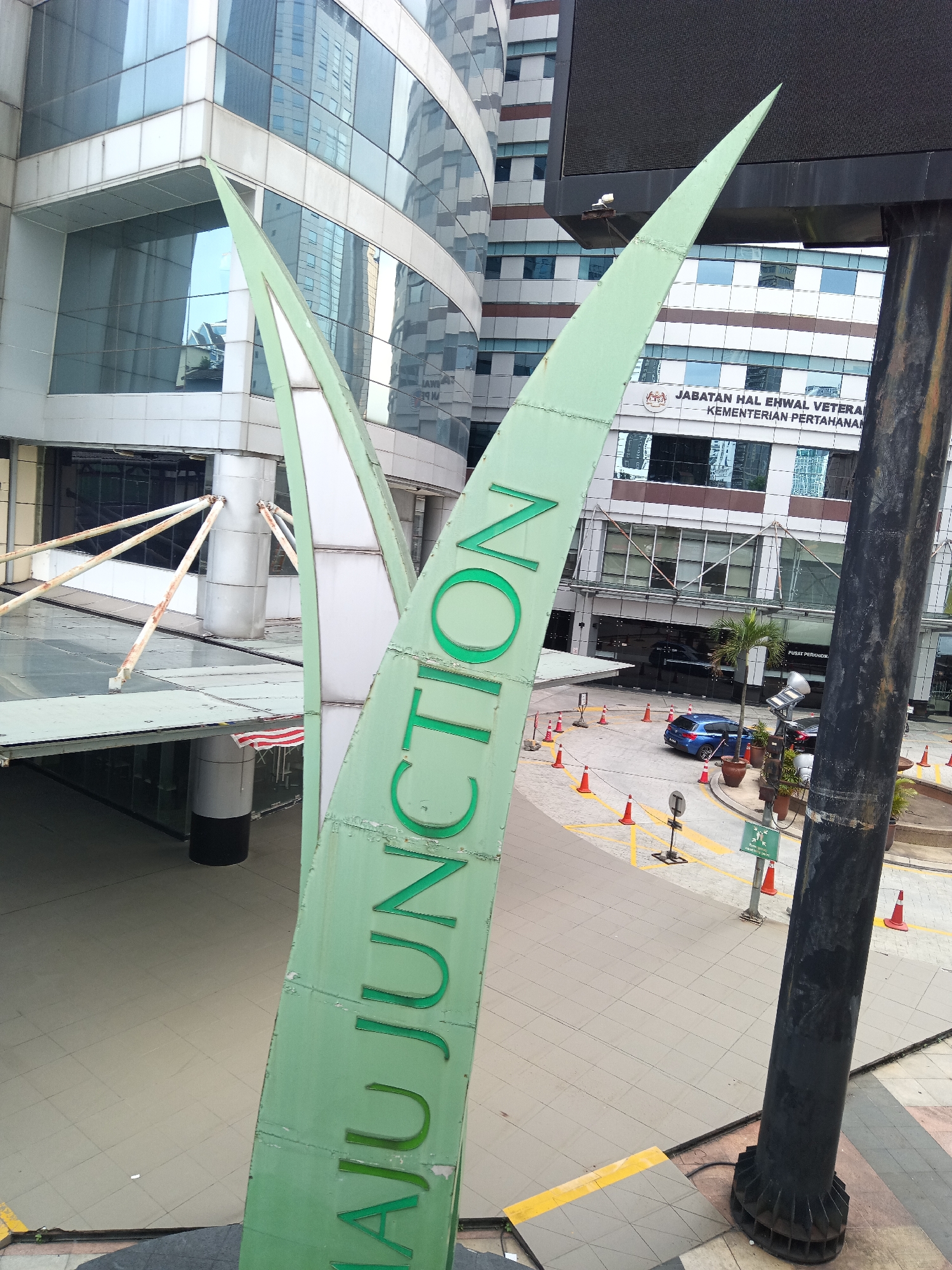 The image shows a view of Maju Junction, a shopping mall located in Kuala Lumpur, Malaysia. In the foreground, a large green sign with the words "MAJU JUNCTION" is visible, angled upwards. Behind the sign, a modern building with reflective glass windows and horizontal banding is seen. Part of a large black pole is on the right side of the frame.
In the middle ground, there is a covered walkway area with pillars and what appears to be an American flag. Further back, a street scene is visible with several orange traffic cones, a blue car, and a palm tree. A building with text in Malay, "JABATAN HAL EHWAL VETERAN KEMENTERIAN PERTAHANAN," is also in the background, indicating the presence of a government ministry building. The overall environment appears to be an urban setting with a mix of commercial and governmental structures.