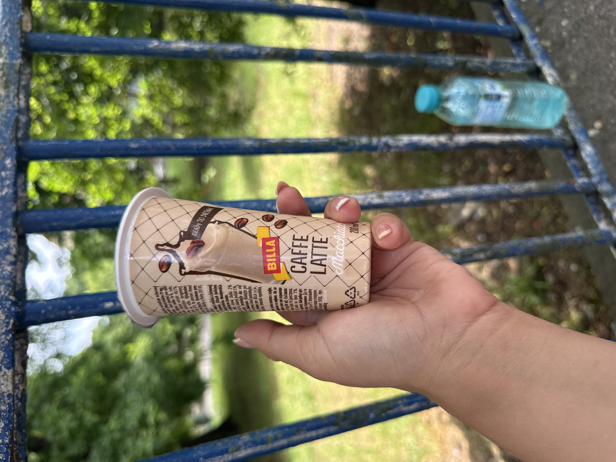 An outdoor, close-up shot shows a person's hand holding a cup of Billa Caffe Latte Macchiato. The cup has a brown and white design with coffee beans and an illustration of the drink. The text on the cup indicates it is "Ready to Drink" and contains 200ml. The person's fingernails are painted pink. In the background, a blue metal fence is visible, and beyond that, out-of-focus green trees and grass suggest a park or outdoor area. A partially visible blue-capped plastic bottle of water rests on the fence. The lighting suggests it is daytime.