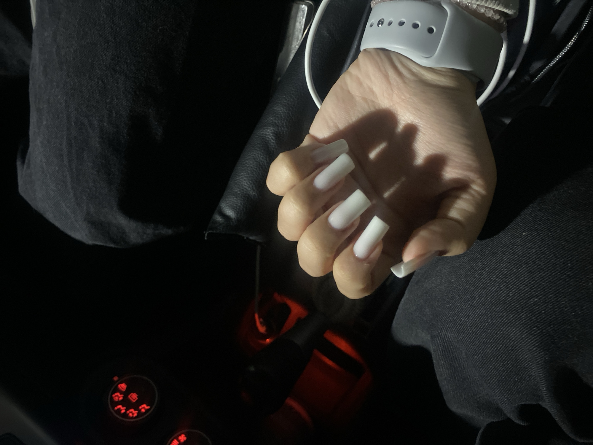 A person with a grey smartwatch and long, white acrylic nails is seated in a car. The person is wearing dark clothing, possibly denim jeans and a jacket, and the car's gear shift and climate control dials are visible. The location, Chaikovsky, Russia, cannot be verified from the image.