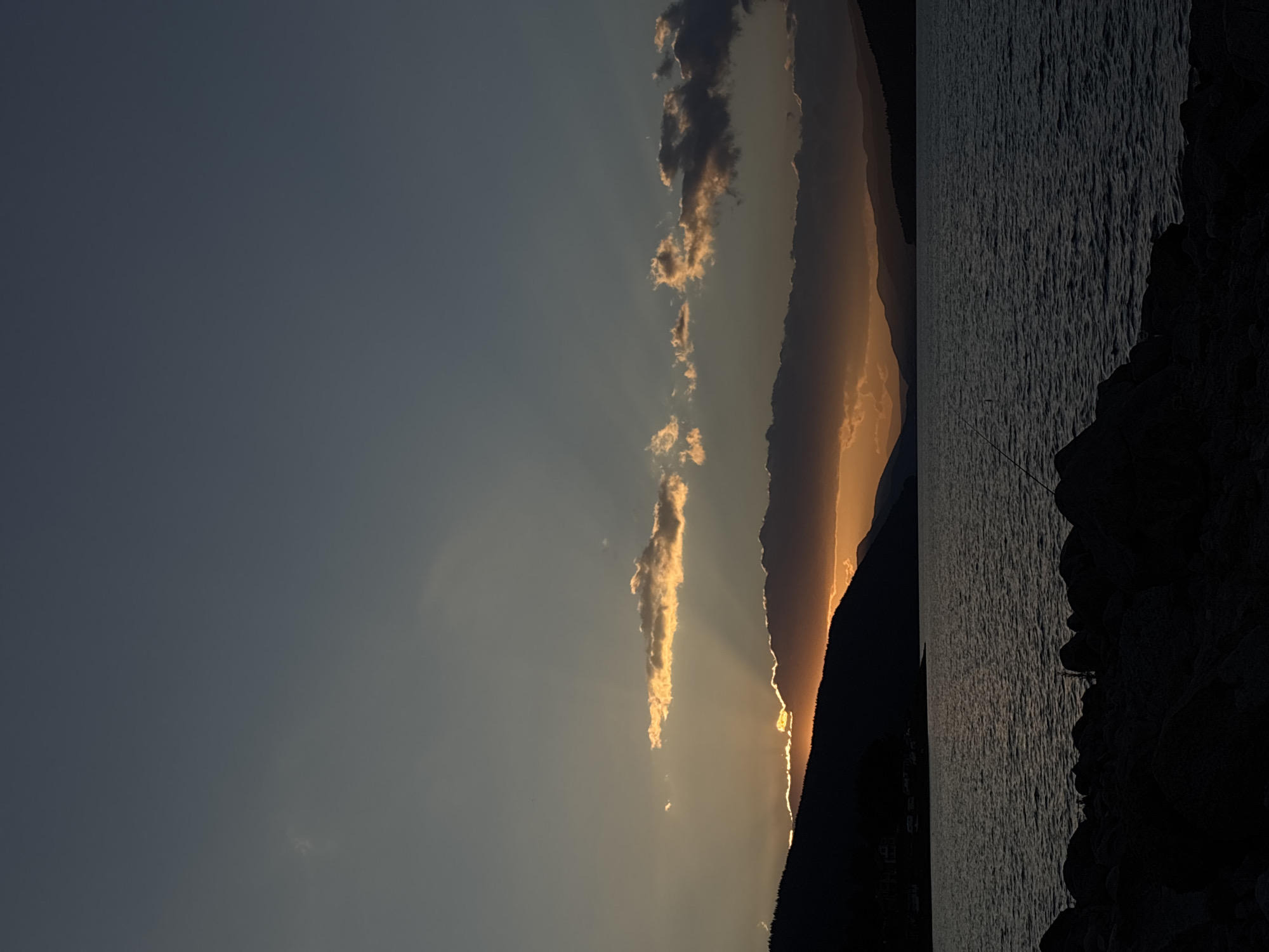A wide shot captures a sunset over a tranquil body of water, likely a lake or bay, bordered by silhouetted hills and mountains. The sky is a gradient of deep blue at the top, transitioning to a muted gray, and then to a warm orange and yellow glow from the setting sun breaking through a band of clouds. Sunbeams fan out from behind a dark cloudbank situated above the horizon. The water's surface is rippled, reflecting the dim light and creating a dark, textured expanse. In the foreground, a rocky shoreline is visible, with large, dark stones creating a rugged edge. A faint, dark line, possibly a fishing rod, is visible in the middle-ground, extending out over the water. The overall atmosphere is serene and peaceful, characteristic of a natural landscape at dusk.