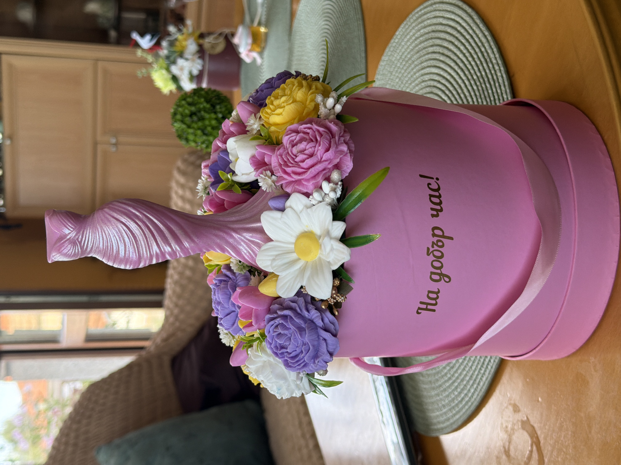 A floral arrangement in a pink cylindrical box is displayed on a wooden table. The box has a pink ribbon handle and text written in gold script on its side that reads "На добър час!" which translates to "Good luck!". The arrangement itself features an assortment of artificial flowers in various colors, including purple, pink, yellow, and white, with a prominent pink, twisted dress-shaped ornament at the center. The background suggests an indoor setting with a window, part of a wicker chair, and wooden cabinetry. The lighting indicates it is daytime. The text on the box is in Bulgarian, a language spoken in Lyubimets, Bulgaria, but the specific location within the city is not discernible from the image alone.