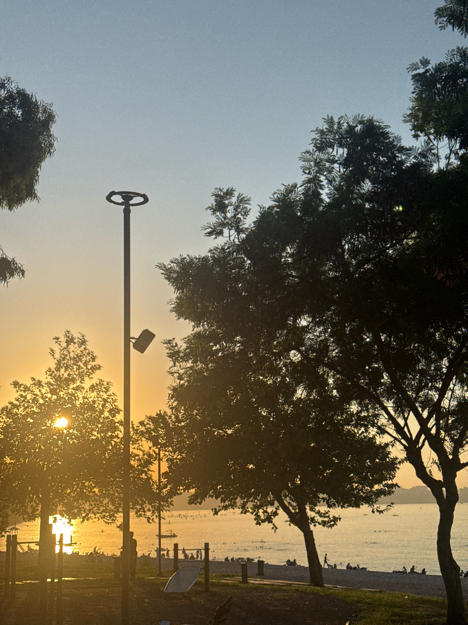 The image depicts a sunset scene on a beach in Antalya, Turkey, likely at Konyaaltı Beach due to the presence of the Taurus Mountains in the distance and the general ambiance of a popular urban beach. The sky is a gradient of pale blue and soft orange, indicative of the golden hour. Silhouettes of trees frame the scene, their leaves catching the warm light. A tall lamppost with a circular fixture at the top stands prominently in the foreground, with a surveillance camera mounted on its side.
On the beach and in the shallow water, numerous people are visible as dark silhouettes. Some are walking along the shore, while others appear to be swimming or wading. A few boats are scattered across the water, and further out, the faint outline of the Taurus Mountains is visible on the horizon. In the immediate foreground, elements of a playground or outdoor gym, including metal bars and a white slide-like structure, are present. The overall atmosphere is one of leisure and relaxation at the end of a day.
