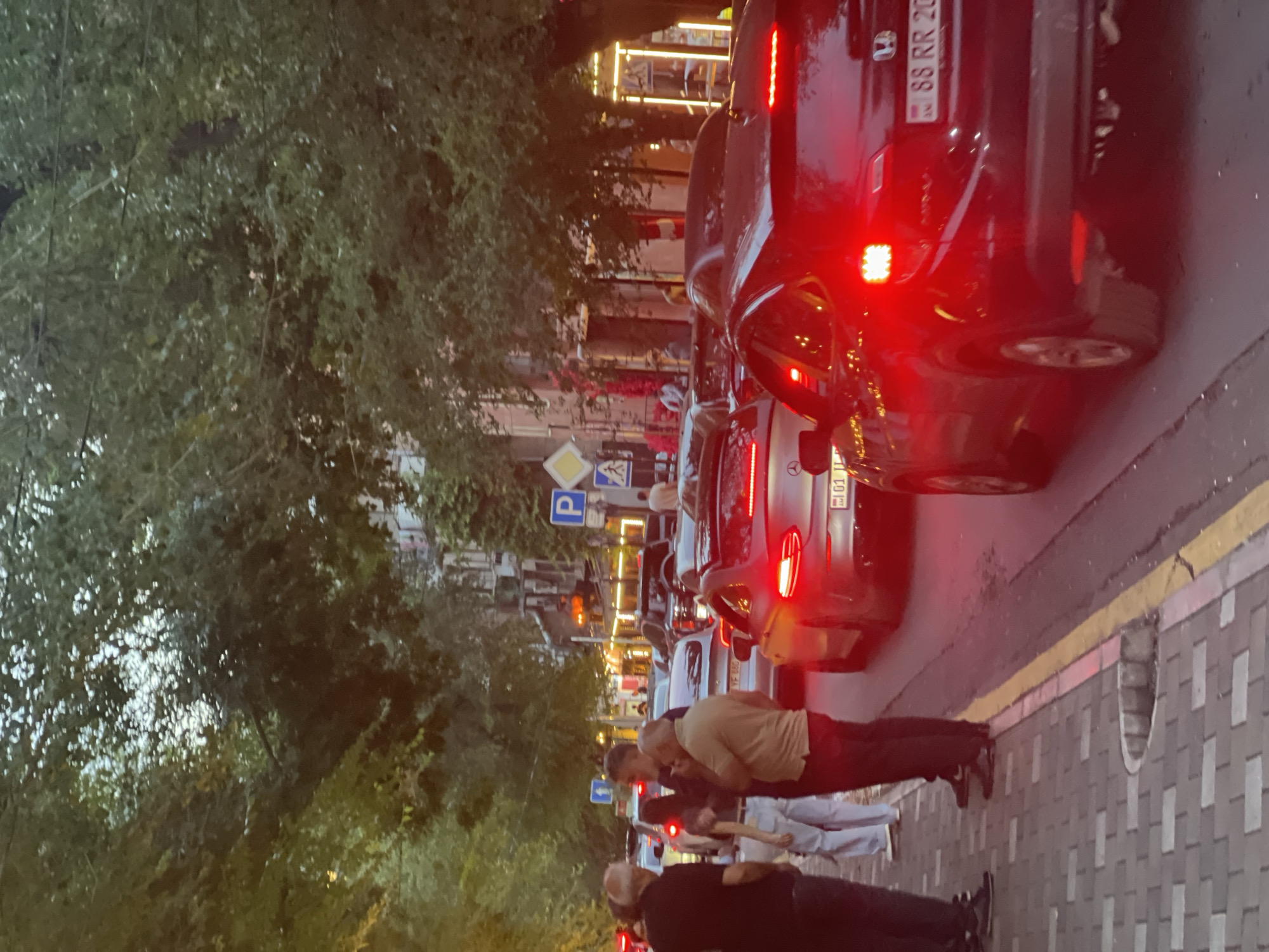 The image captures a street scene in Yerevan, Armenia, likely during the evening hours, given the artificial lighting and the twilight sky. The foreground is dominated by a traffic jam, with several cars lined up on the asphalt road. The car closest to the viewer, a dark-colored sedan, has its red brake lights illuminated, indicating it is stopped. Another silver car in front of it also has its brake lights on. A third silver car, further down the lane, displays a license plate that appears to be from Armenia.
On the left side of the image, a sidewalk is visible, paved with interlocking bricks. Several people are standing on the sidewalk, conversing. They are dressed in casual clothing. One man in a black shirt and dark pants stands facing away from the camera. To his right, a woman in a light-colored top and white pants is also present. Further to the right, a man in a tan shirt and dark pants is engaged in conversation with another man.
The street is lined with trees, their branches extending over the road, creating a canopy effect. Buildings can be seen in the background, some with lit windows and illuminated facades. Traffic signs, including a blue parking sign with a white 'P', are visible on poles along the street. The overall atmosphere suggests a busy urban environment experiencing a traffic delay.