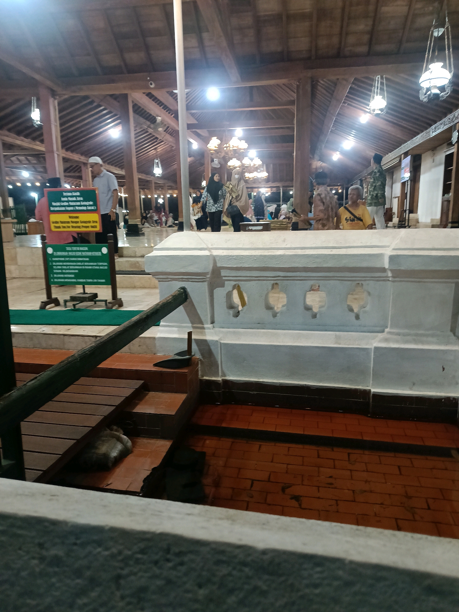 The image captures an outdoor scene at night, likely a religious or historical site in Yogyakarta, Indonesia, given the architectural style and the presence of a tomb. A stone tomb with decorative carvings sits in the foreground, with a wooden railing in front of it. To the left, a green sign with text in Indonesian and English provides regulations. Several people are visible in the background, some in traditional Islamic attire, suggesting a mosque or a shrine. The atmosphere is illuminated by hanging lamps, creating a warm, ambient light. Wooden structures and roofing are prominent, indicating traditional Indonesian architecture. A cat is visible near a water feature in the lower left of the frame.