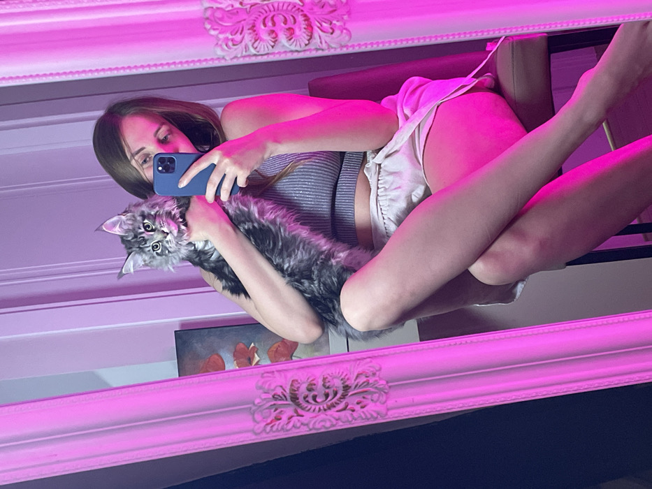 A mirror selfie shows a woman in a strappy top and shorts holding a gray Maine Coon cat. The woman holds a blue smartphone and looks at its screen. The cat looks directly at the viewer. The mirror has an ornate, white frame illuminated by a pink light. A painting with red poppies is partially visible in the reflection. A chair back is behind the woman.