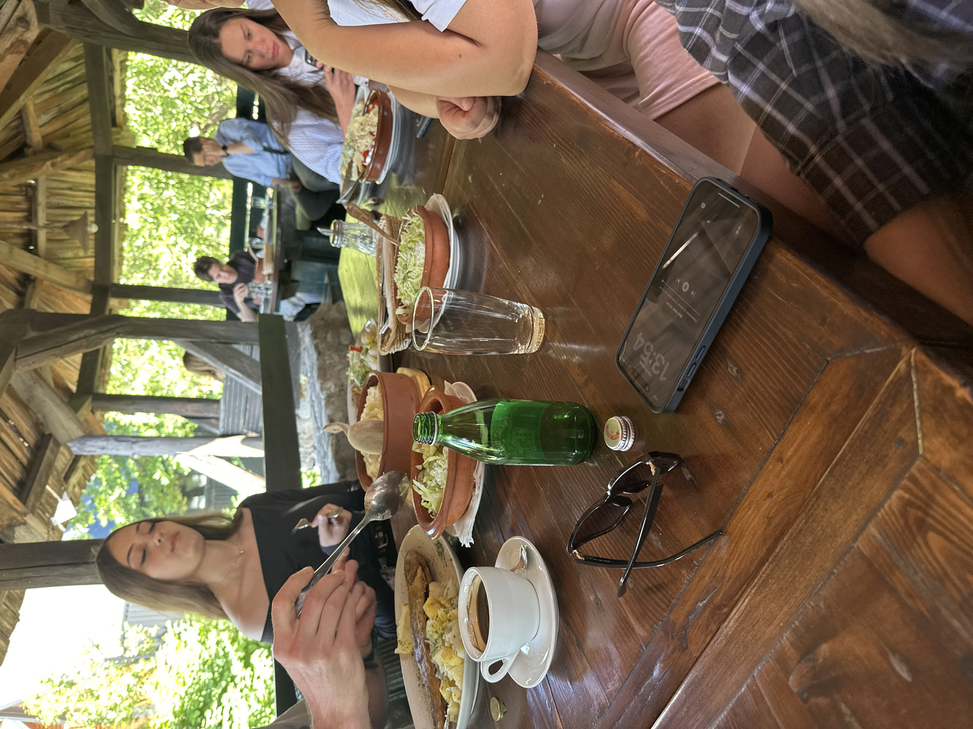 A group of people are dining at an outdoor restaurant in Kolašin, Montenegro, under a wooden-beamed canopy. On a wooden table, there are several dishes of food, including a plate of fish with potatoes and a bowl of salad. A cup of coffee sits next to a green glass bottle and a smartphone. Sunglasses are placed on the table, and a few bottle caps are scattered around. In the background, more diners are visible, along with lush green trees and vegetation.