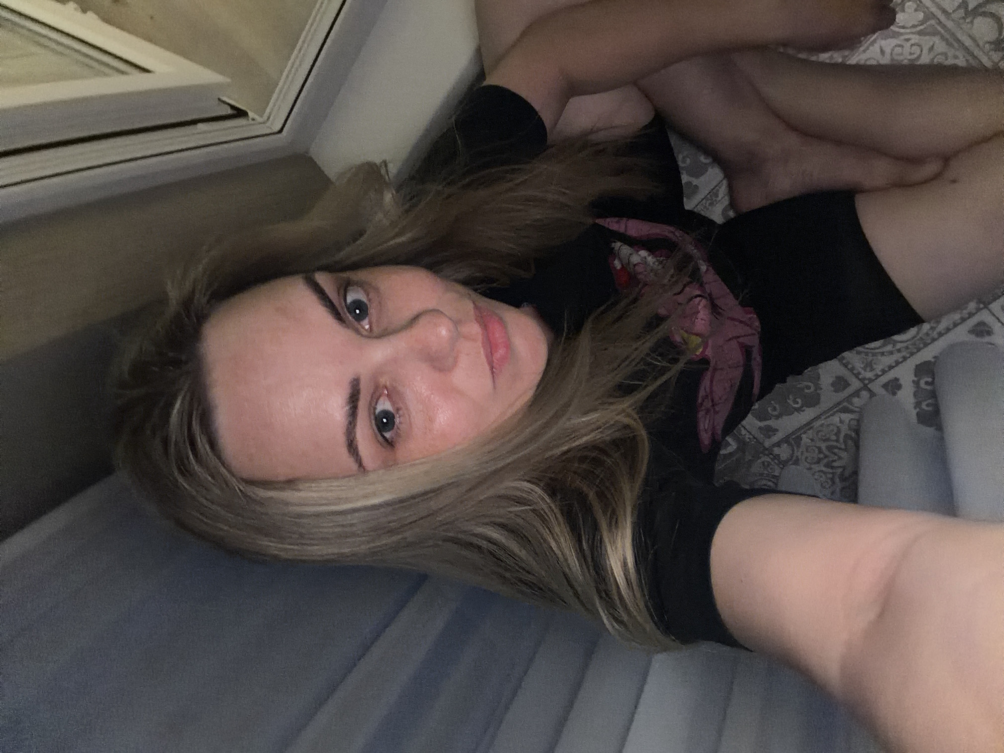 A woman with blonde hair is lying on a gray surface, possibly a bed or mattress. She is wearing a black shirt and black shorts. Her legs are crossed. A window is visible in the upper left corner. The background includes patterned bedding. It is impossible to determine the location as Votkinsk, Russia or any other specific location from the image alone.