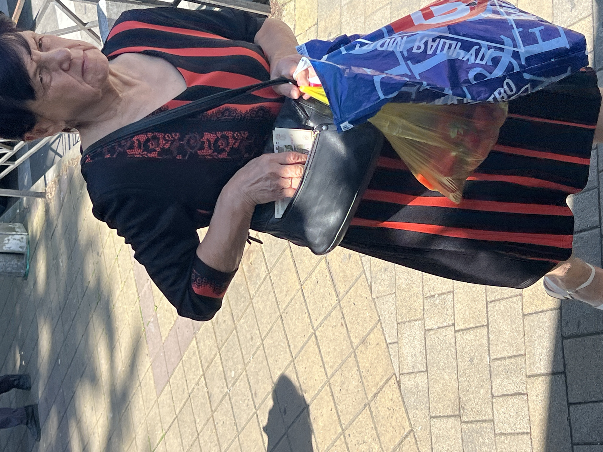 An older woman, likely in her 60s or 70s, stands on a paved surface, probably a sidewalk, under bright sunlight, indicated by distinct shadows. She has dark hair with bangs and a neutral facial expression.
She is dressed in a black dress with prominent horizontal red stripes and intricate black lace detailing over a red background on the bodice and sleeve cuffs. Her right foot, visible at the bottom right, wears a white sandal.
The woman is holding a black handbag or clutch with her left hand, where several banknotes are visible, suggesting she is either withdrawing or depositing money. Attached to or placed beside her handbag is a large blue and white reusable shopping bag. Inside this blue bag, a clear yellow plastic bag holds several red items, possibly fruits like apples or vegetables such as tomatoes.
Visible text on the blue shopping bag includes the Russian phrase "ЛУЧШАЯ ЦЕНА" (BEST PRICE) and fragmented letters like "BO". The banknotes have small, intricate designs but no clearly legible denominations.
The setting appears to be an urban outdoor environment in Sochi, Russia, with light-colored square pavers forming the ground and a metallic structure, possibly a fence or railing, in the blurred background. The overall scene captures a moment of daily activity, likely shopping or handling personal finances, in a sunny outdoor locale.
