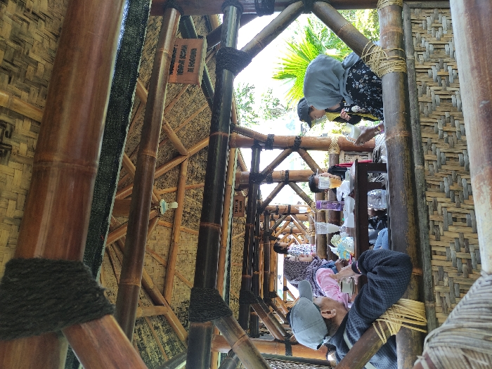 The scene takes place in an open-air bamboo structure located in the Kecamatan Pujon district of Indonesia, near the geographic coordinates -7.854 latitude, 112.453 longitude. The structure is constructed from woven bamboo, and a woven bamboo roof provides overhead cover. There are five people present, seated at a table in the center of the bamboo structure. The table is wooden and covered in a white tablecloth. Two adults and two children are seated at the table, with a fifth person seated at a table off-camera, to the right. The adults appear to be engaged in conversation, while the children appear to be eating. There is a variety of food and drink on the table, including a plate of food, a glass of what appears to be iced tea, and a glass of water. The scene is bathed in warm, natural light, and the atmosphere is relaxed and peaceful.