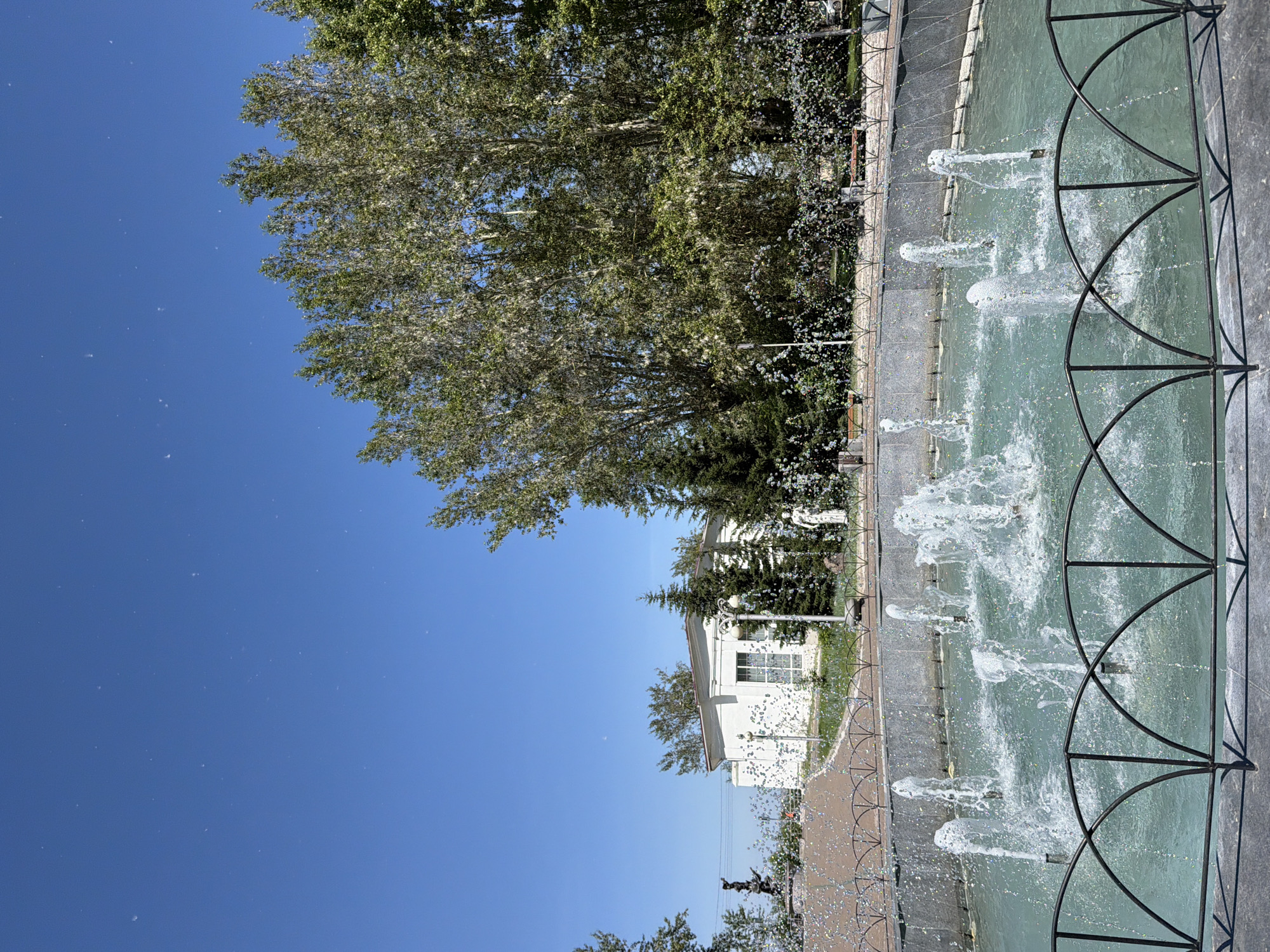The image shows a fountain in a park with trees and a white building in the background. The fountain is circular with water spraying from multiple jets. The water is a light blue-green color. There are trees with green leaves behind the fountain, and a white building with windows and a red roof. The sky is clear and blue with some small white specks that appear to be pollen or seeds. The scene appears to be in an urban park setting.