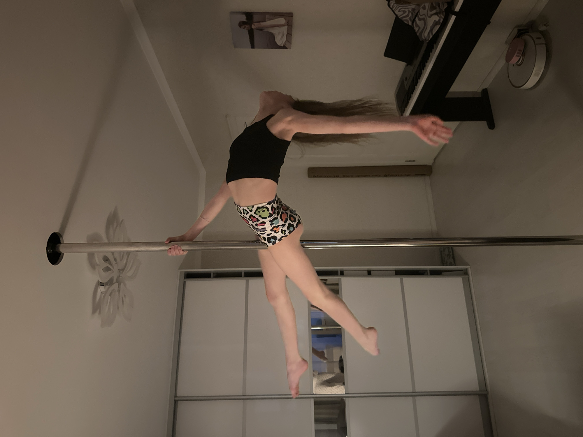A young woman is suspended from a horizontal bar, seemingly performing a gymnastic or acrobatic maneuver in what appears to be a domestic setting. She wears a black top and animal print shorts. Her hair is long and flows outward. She is holding the bar with her left hand while her right arm is extended to the side. She has one leg extended, and the other is bent. The location seems to be indoors. Visible in the background are a wall-mounted light fixture, a wardrobe, a piano, and what appears to be a picture on the wall. No specific location information is included.