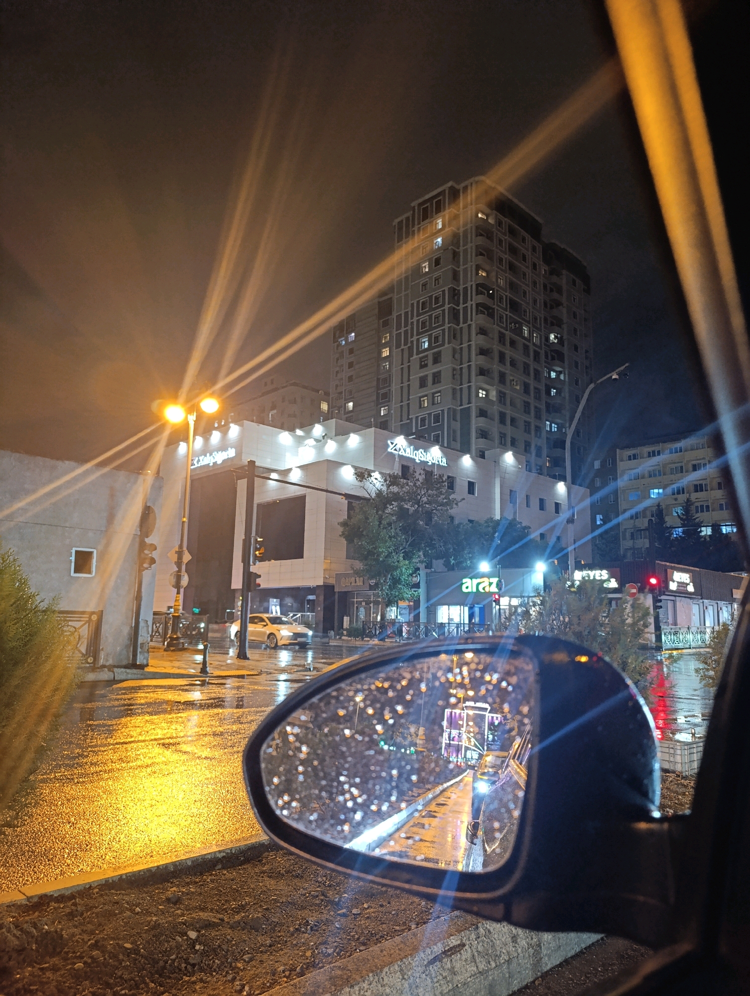 The image captures a night scene in Baku, Azerbaijan, likely on a rainy evening. The perspective is from inside a vehicle, looking out the side window and into the side-view mirror. The wet asphalt reflects the bright lights of the city, creating a shimmering effect. In the background, tall buildings with illuminated windows stand against the dark sky. A prominent street lamp with two bright lights illuminates the wet street and a nearby building with signage. The side-view mirror reflects the road behind the vehicle, showing more of the wet street and lights, with raindrops visible on the mirror's surface. There are no visible people in the scene. The overall atmosphere is urban and nocturnal, with the rain contributing to the reflective and moody ambiance.