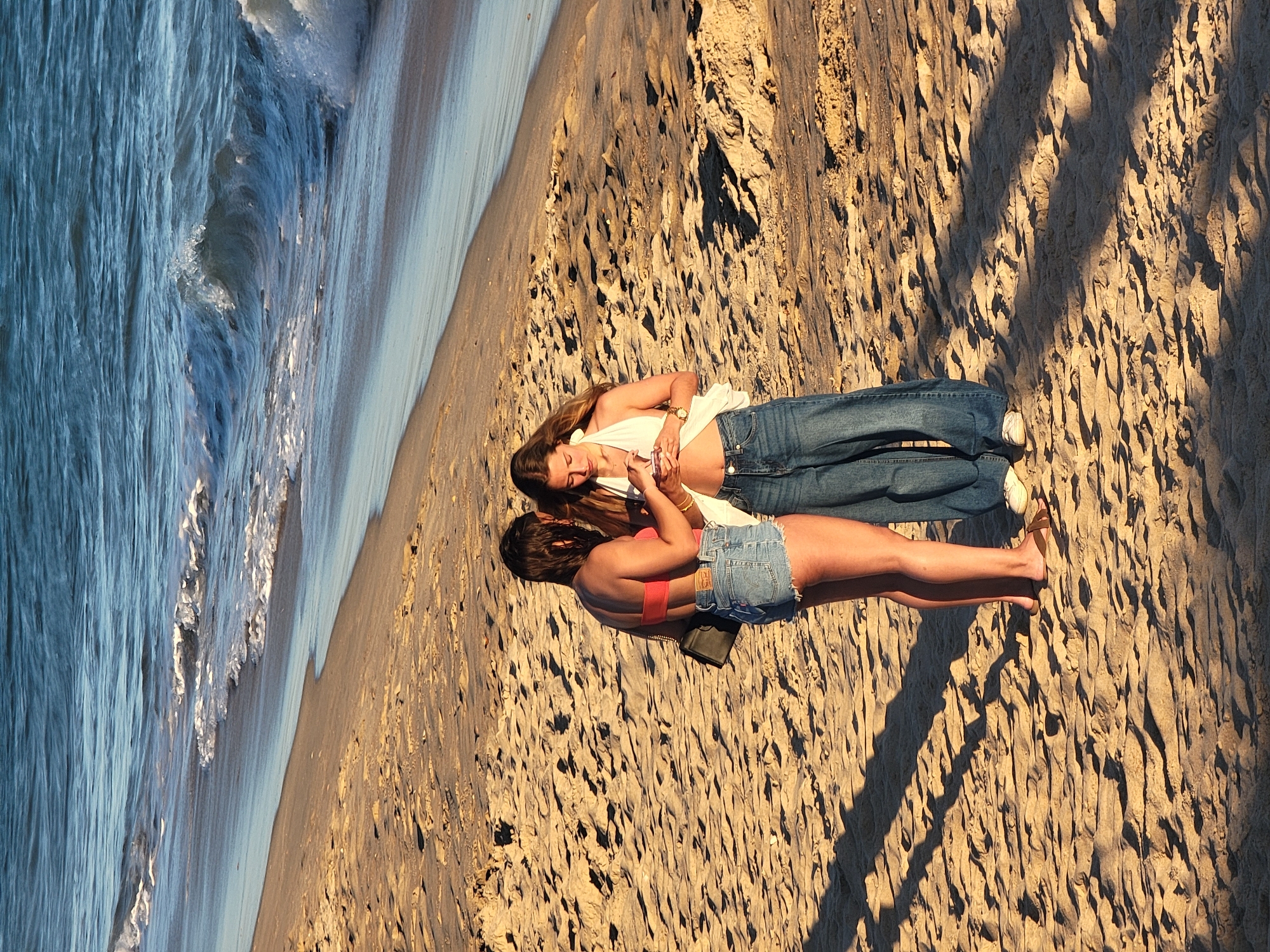 Two young women stand on a sandy beach in Quarteira, Portugal. The woman on the left wears a coral-colored bikini top and denim shorts. The woman on the right wears a white halter top and loose-fitting blue jeans. Both are looking down at a phone, with the woman on the right holding it. The ocean with gentle waves is visible in the background. The sand is textured with shadows cast from an unseen object, likely dunes or vegetation. The sun appears to be low in the sky, casting a warm glow on the scene.