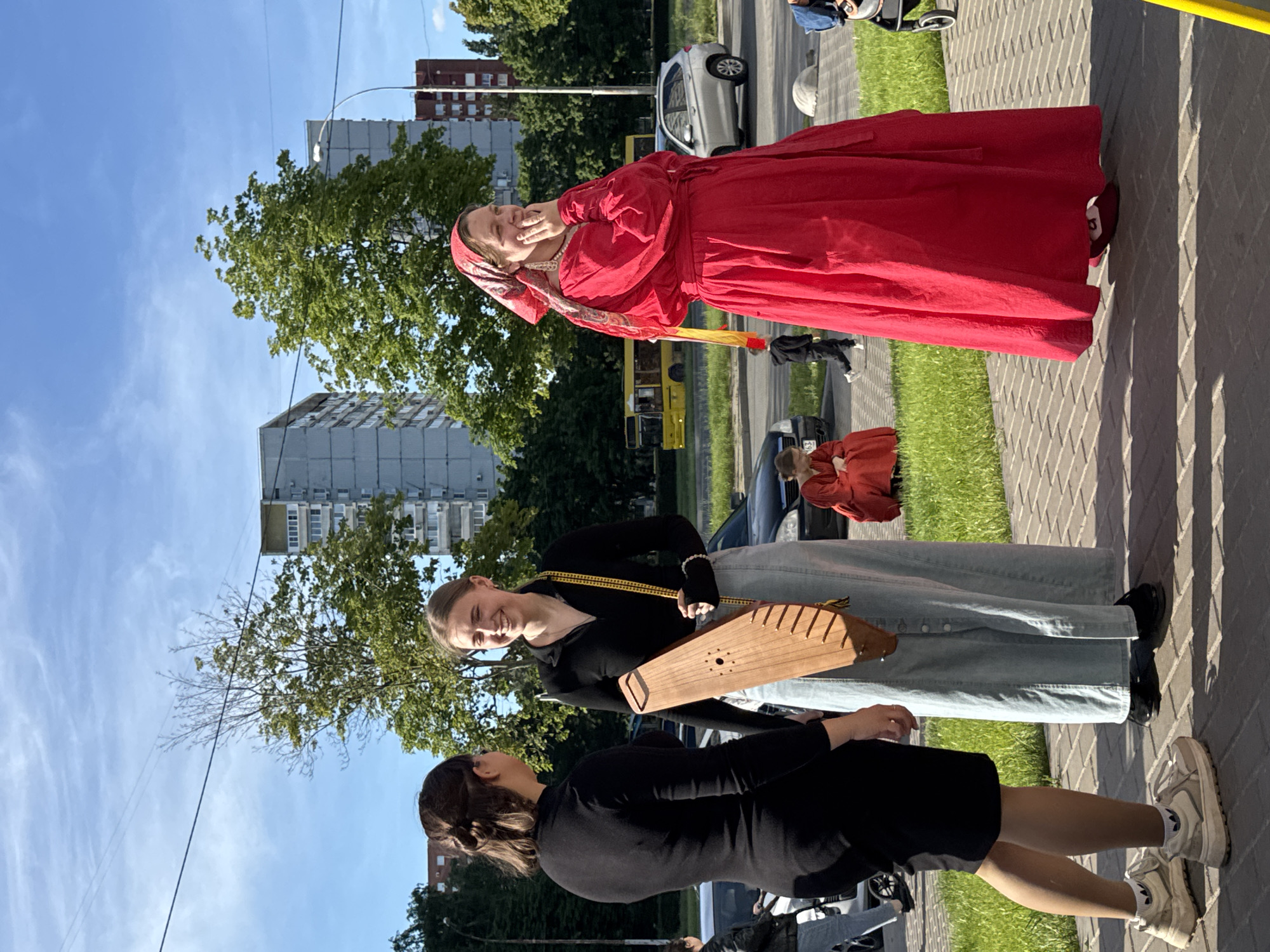 This image captures three women outdoors on a bright, sunny day, standing on a paved sidewalk next to a street. The photograph is oriented vertically, with the subjects appearing to stand upright.
The most prominent figure is an older woman on the right, dressed in a striking, long-sleeved red traditional dress. She wears a patterned red, white, and gold headscarf and a white pearl necklace. Her hands are near her mouth, and she is smiling widely, looking up and to her right, conveying a sense of joy or amusement. Red shoes are visible beneath her dress.
To her left stands a younger woman with light hair, also smiling. She wears a black long-sleeved top and a light blue, button-front midi skirt made of denim-like material. She is holding a distinctive, light-brown wooden stringed instrument, resembling a gusli or kantele, which hangs from a yellow strap over her shoulder.
Further to the left, a third younger woman with dark hair is partially visible from behind and the side. She wears a black dress or top and skirt combination, along with light-colored sneakers and white socks. She is looking towards the woman holding the instrument.
The setting is an urban or suburban environment, featuring a paved sidewalk, a narrow strip of grass, and an asphalt road with parked cars (a silver sedan and a darker vehicle, with a yellow bus further down the street). In the background, there are multi-story residential buildings, some grey and some reddish-brown, along with several green trees. The sky is clear and blue with minimal clouds, and strong shadows on the pavement suggest bright sunlight, indicating midday or early afternoon. The overall atmosphere appears cheerful and relaxed. No visible text is legible in the image.