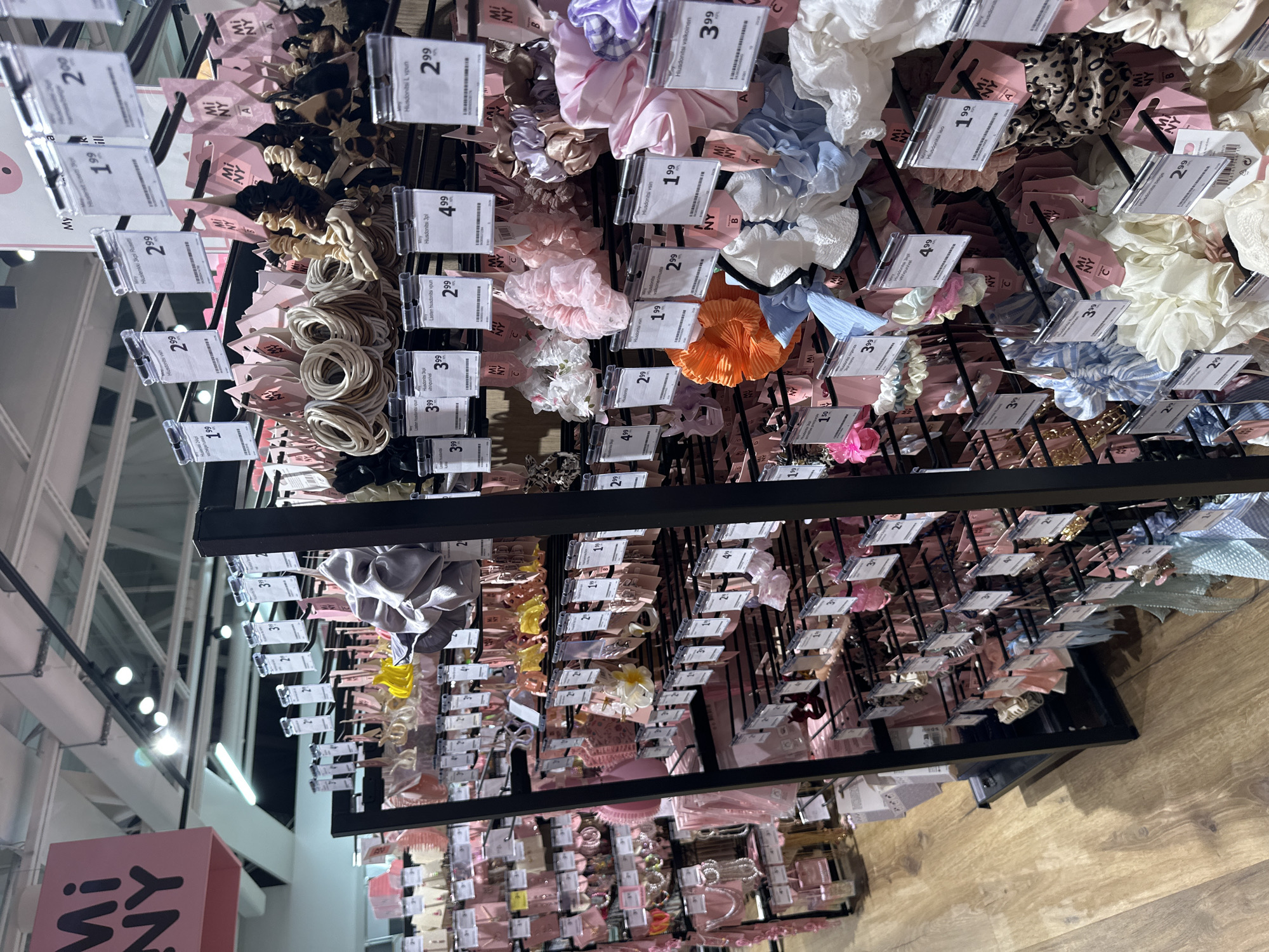 The image shows an aisle in a retail store selling hair accessories. The shelves are stocked with various items such as scrunchies, hair ties, and clips, predominantly in pastel colors like pink, blue, and white, as well as some leopard print and floral patterns. Many items are displayed on black metal racks, with price tags and product information cards attached. A prominent pink sign with "Mi NY" written in black letters is visible in the upper left corner, indicating the brand or store name. The flooring appears to be made of light-colored wood planks. The lighting is bright, typical of a retail environment. There are no people visible in the image. The scene is a close-up view of the merchandise display.