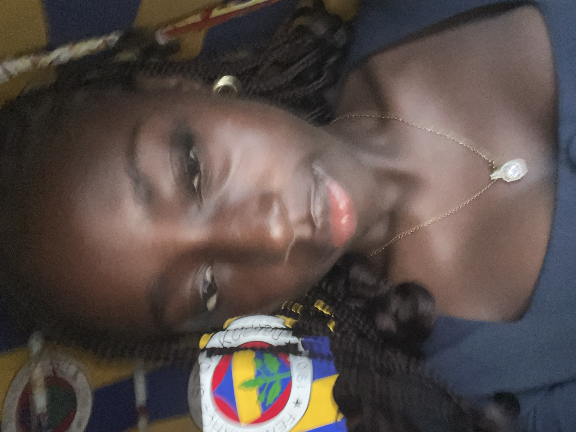 A dark-skinned person with braided hair is lying on a bed with a Fenerbahçe Spor Kulübü can or mug. The person wears a gold necklace and earring. The location is potentially in Galadimawa, Nigeria.
