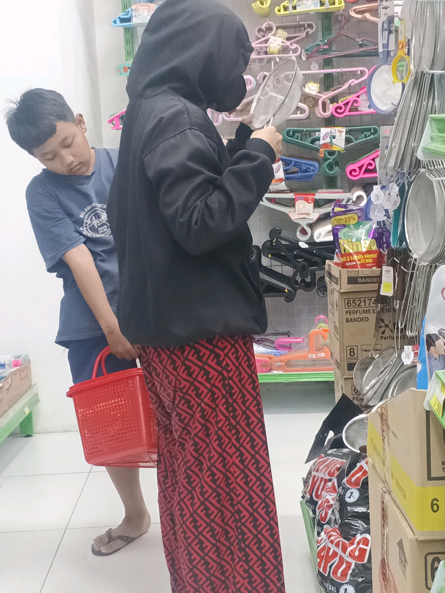 In Kemangsen, Indonesia, a young boy wearing a blue t-shirt stands in front of shelves stocked with household goods. To his right, a person wearing a black hooded jacket and red patterned pants holds a metal sieve. The boy appears to be looking down at the floor while the person in the hooded jacket is examining the sieve. A red plastic basket is held by the person in the hooded jacket, and the boy's right hand is close to it. The background displays various items on shelves, including hangers, plastic containers, and packaged goods. The floor is tiled.