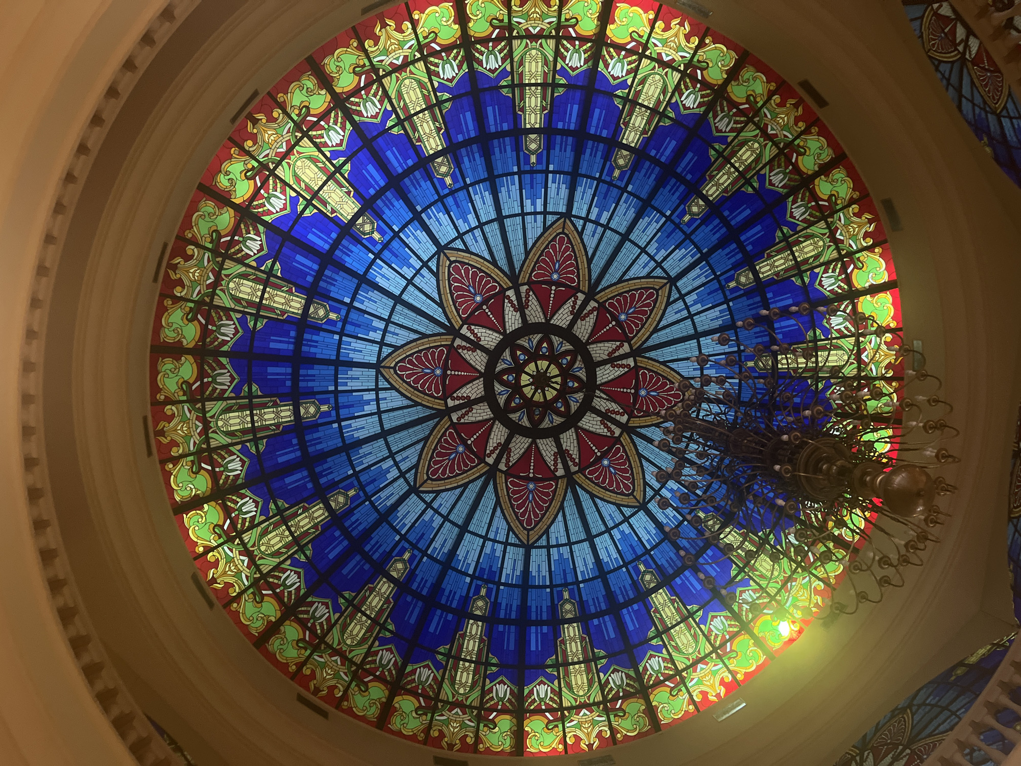This is a low-angle shot looking up at a large, ornate stained-glass dome ceiling with a chandelier hanging in the foreground. The dome features intricate geometric patterns in shades of blue, red, green, and yellow, with a central floral motif. The surrounding architecture is a warm, golden-brown color, with decorative molding along the curve of the ceiling. The chandelier appears to be made of brass and has multiple arms with candle-like fixtures. The lighting is somewhat dim, with a noticeable lens flare in the upper left quadrant. The scene appears to be indoors, possibly in a public building such as a theater, museum, or hotel. The image does not contain any people or animals.