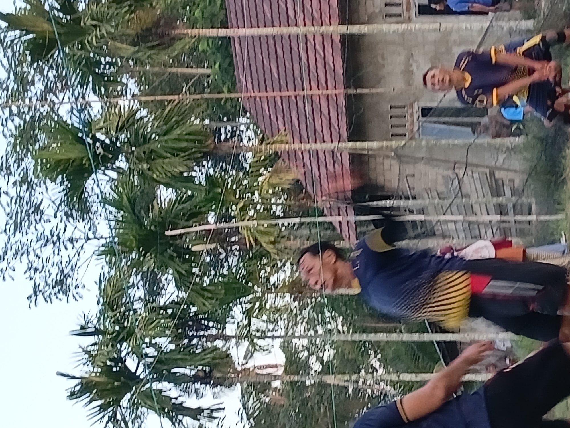 In Margo Rukun, Indonesia, an outdoor scene unfolds with a group of individuals engaged in what appears to be a recreational activity, possibly volleyball. In the foreground, a man in a blue and yellow athletic shirt and black shorts is in mid-action, looking upwards with his mouth open, suggesting he is about to hit or receive a ball. His body is crouched, indicating a dynamic movement. To his left, another person, only partially visible from the waist down, wears black shorts and a blue shirt, with their left hand extended forward. In the background, several tall palm trees with slender trunks and green fronds tower over the scene. A building with a reddish-brown tiled roof and pale, textured walls is visible behind the trees. The building has several windows and a generally weathered appearance. Further in the background, another individual in a blue shirt and dark shorts is also observed. The overall setting suggests a casual, perhaps community-based gathering in a tropical or sub-tropical environment.