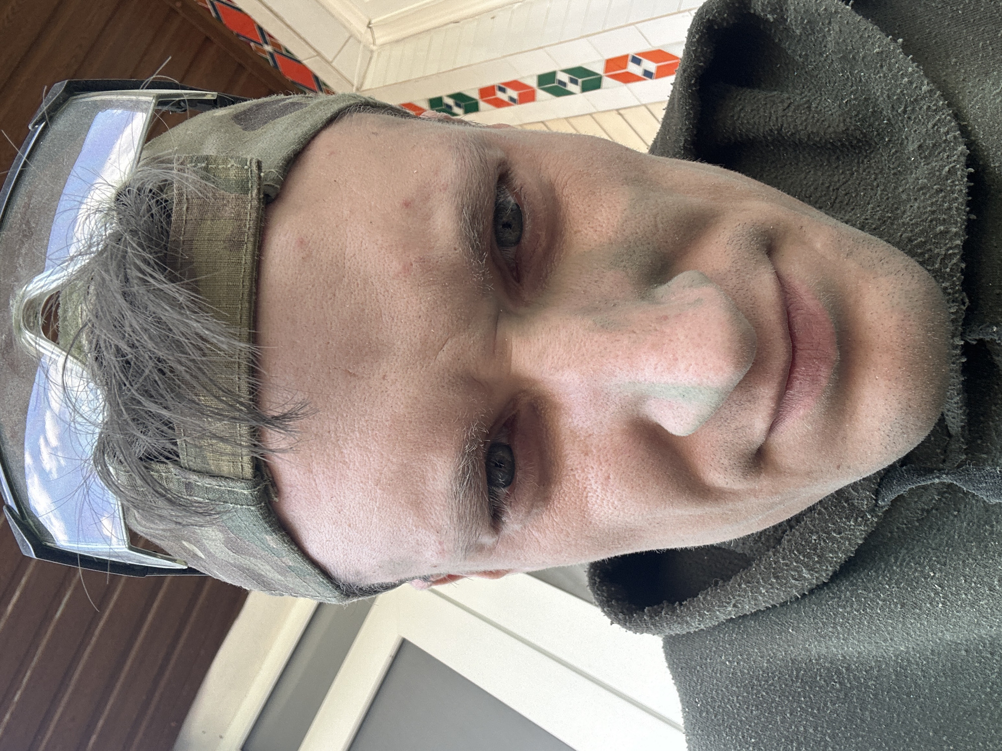 The media file is a close-up, low-angle, portrait-style photograph of a person's face, likely taken by the subject themselves. The person is wearing a camouflage baseball cap and a dark, possibly green, hooded garment that appears somewhat worn. There are smudges of what seems to be dirt or dust on their face, particularly on their nose and cheeks. The lighting suggests it might be daytime, possibly outdoors. The background shows a wooden structure and a tiled pattern with orange, green, and white squares, possibly on a porch or exterior wall. The subject's expression is neutral, and their gaze is directed slightly away from the camera. The overall impression is of someone engaged in an outdoor activity or manual labor. The location context is Makiv, Ukraine, but no specific landmarks are clearly identifiable in the image to confirm this. There is no visible text in the image.