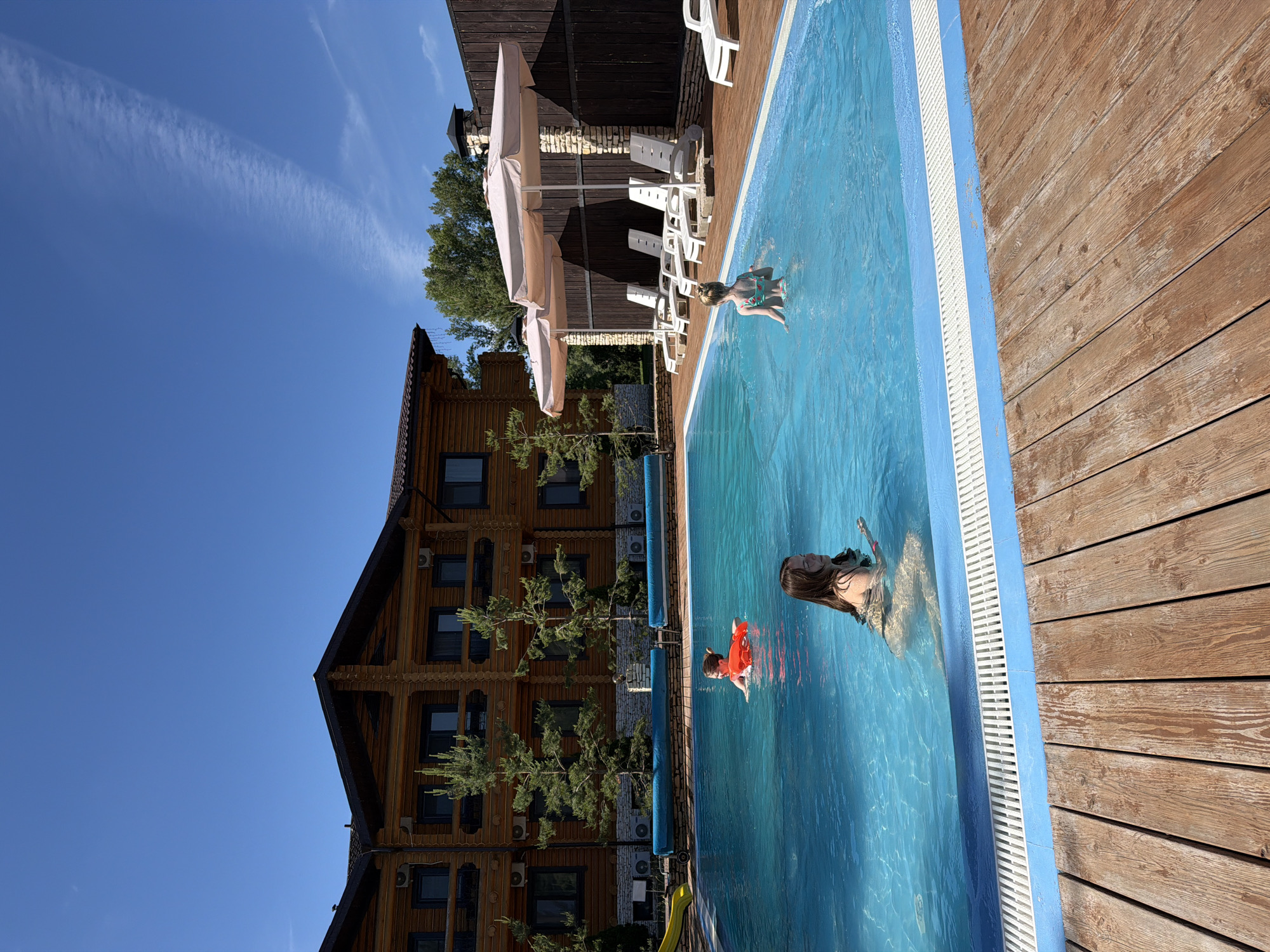 In Красный Ключ, Russia, a picturesque scene unfolds at an outdoor swimming pool. The pool, with its crystal-clear blue water, is surrounded by a wooden deck. In the foreground, the warm tones of the weathered wood planks draw the viewer into the scene.
Three individuals are enjoying the water. In the middle of the pool, a young girl with blonde hair pulled back, wearing a green and pink patterned swimsuit, is facing away from the camera. She is splashing in the water, with ripples emanating from her presence. Closer to the foreground, a woman with long dark hair, wearing a dark-colored swimsuit, is partially submerged in the water. She is looking to her right and holding a small object in her hand. Further back, near the middle of the pool, another individual, whose face is not clearly visible, is wearing an orange flotation device.
The pool area is framed by a large, multi-story wooden building with numerous windows and balconies, suggesting it might be a hotel or resort. Two large beige umbrellas provide shade over a row of white lounge chairs situated on the wooden deck. Beyond the pool and lounge area, a wooden fence encloses the property, and lush green trees are visible in the background. The sky above is a vibrant, clear blue, with wisps of white clouds stretching across it. The scene is bathed in bright sunlight, indicative of a warm, summer day.