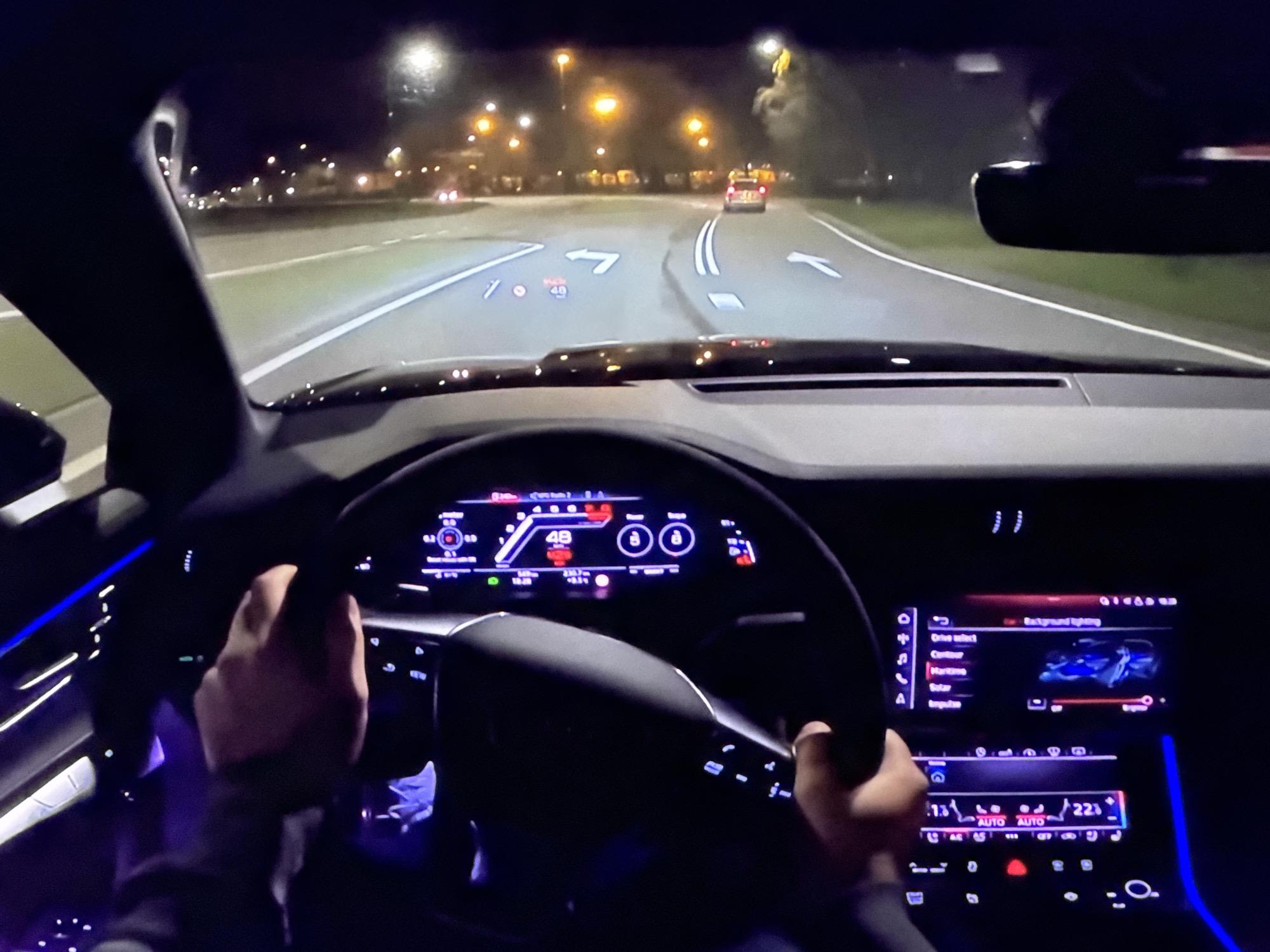 The image shows a first-person view from the driver's seat of a car at night. The car is driving on a road with streetlights and trees visible in the background. The dashboard displays various information on a digital screen, including speed (48 km/h), navigation arrows, and other vehicle data. The steering wheel is visible, with a person's hands on it. The car's interior is illuminated by ambient lighting. There are no discernible landmarks that would indicate the location of Bezden, Bulgaria.