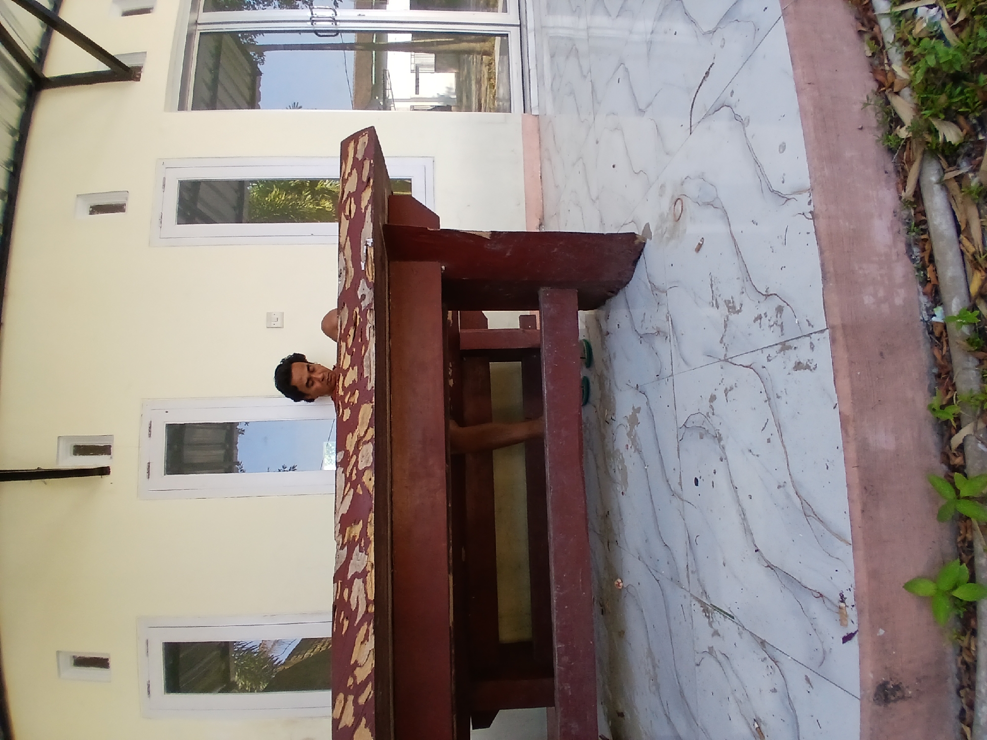A man is sitting at a wooden table, his head down as if he is working or reading. The table is large and appears to be made of dark red wood with a lighter wood inlay on the top. The man is wearing a dark shirt and has dark hair. He is positioned on a tiled patio with white tiles. In the background, there is a building with yellow walls and windows. To the right of the building, there is a large glass sliding door. The overall scene suggests an outdoor setting, possibly a home or a communal area, in Pejaten, Indonesia.