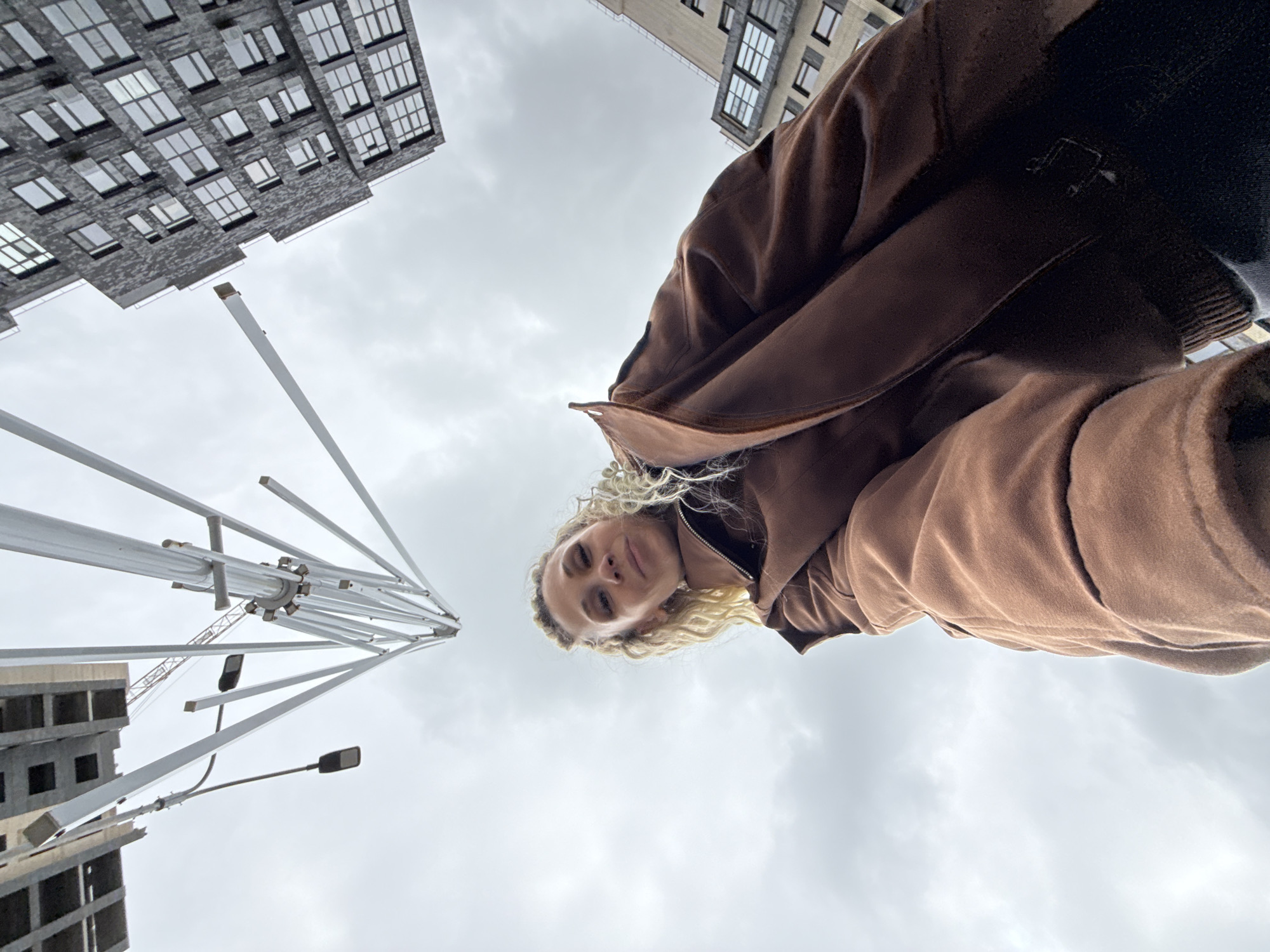 A low-angle, wide shot captures a woman outdoors with urban surroundings. The woman, with light blonde curly hair, wears a brown jacket and looks towards the camera. Behind her, two high-rise buildings flank a cloudy sky. To the left, a metal structure with multiple poles extends upward. A crane is visible near the base of the structure, and streetlights are mounted on one of the poles. Given the information provided, the location is likely Rusanovo, Russia.