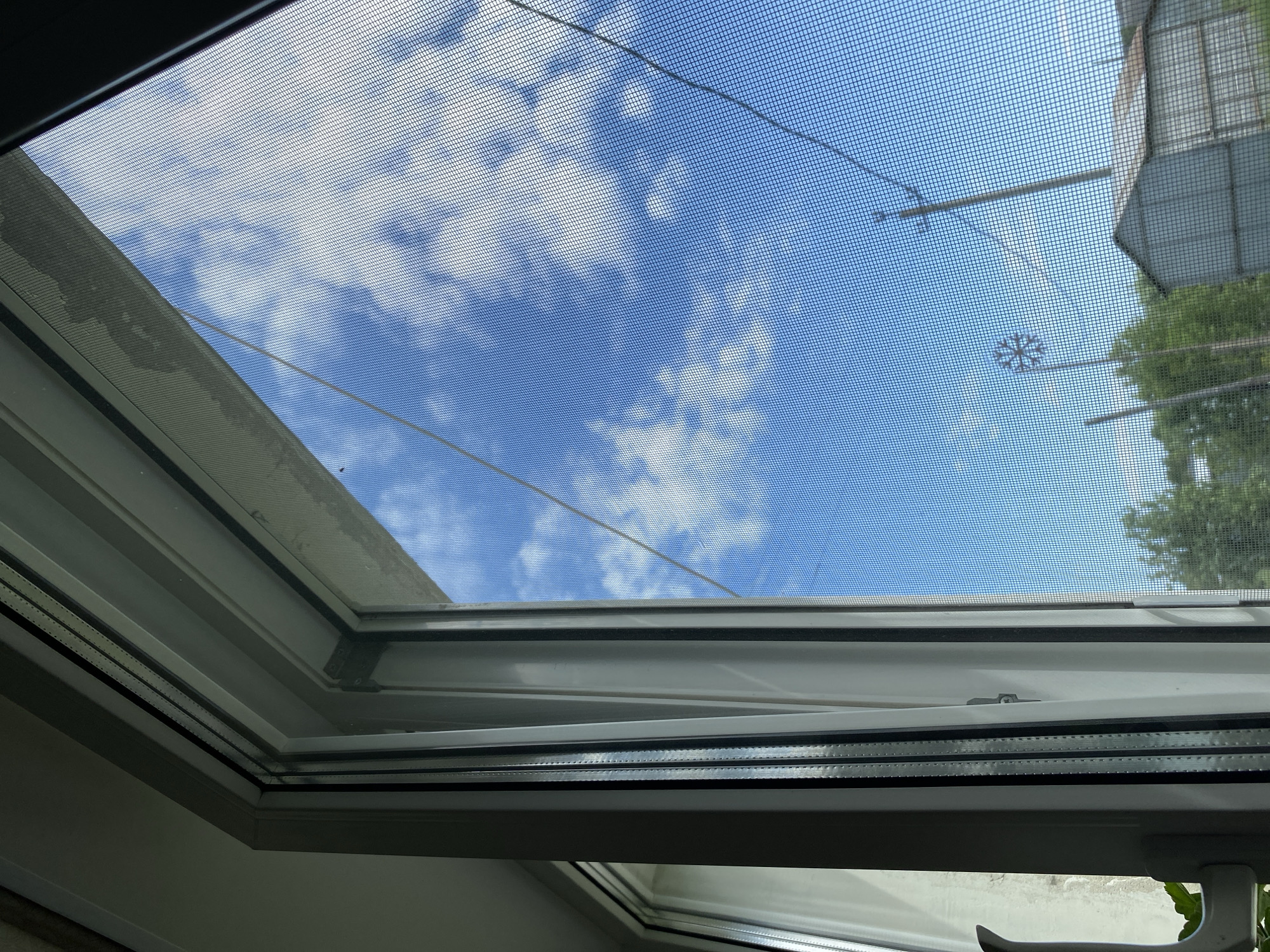 The image shows an open skylight with a screen. Through the screen, a bright blue sky with puffy white clouds is visible. Electrical wires are strung across the view of the sky, and a nearby utility pole or structure is partially visible on the right side. The frame of the skylight is white and grey, and the interior of the room is also visible.