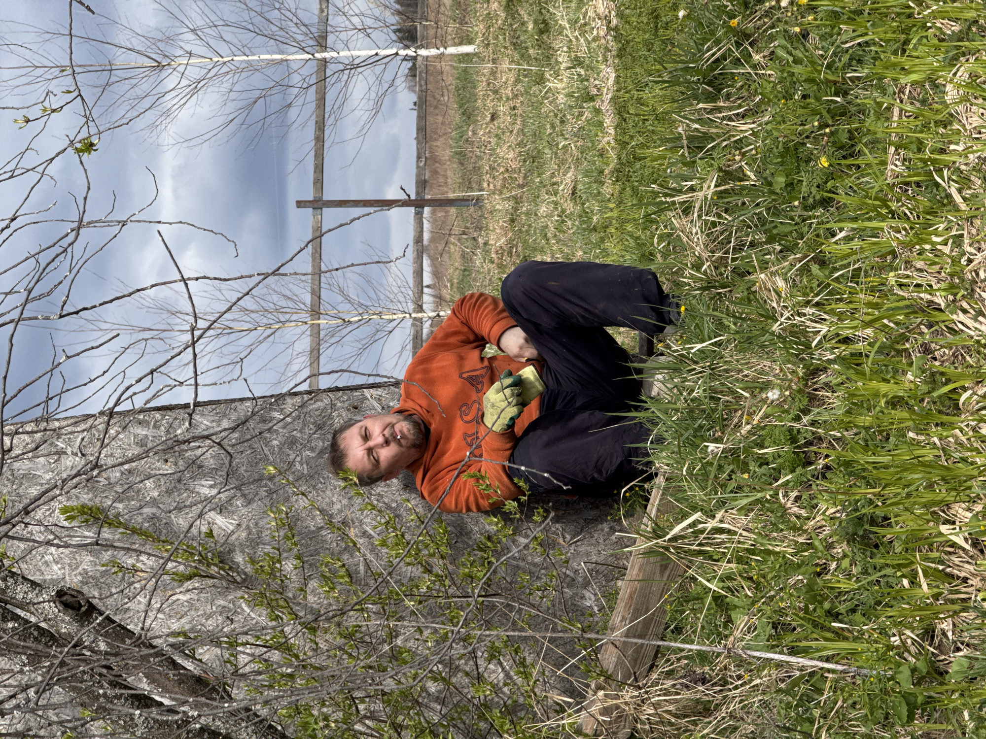 A middle-aged man is lying on a sheet of plywood propped against a fence in a grassy field. He is wearing an orange sweatshirt, dark pants, and gloves. He appears to be smoking. The scene is outdoors, possibly near the city of Усть-Тунтор, Russia, based on geographic context. The background includes a fence, trees, and a field.