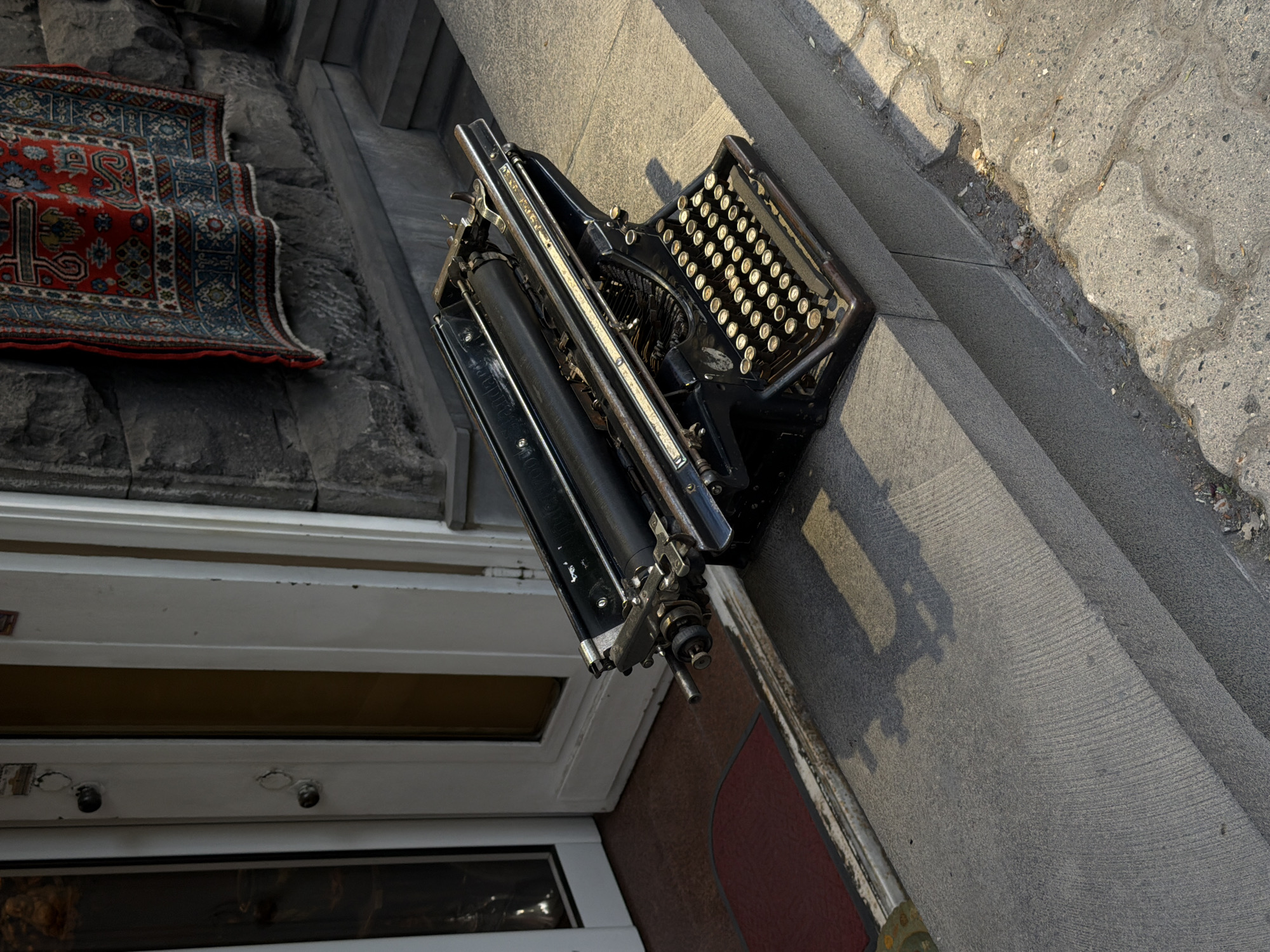 This outdoor, high-angle shot captures a vignette of antique items on stone steps, likely at a building entrance in Yerevan, Armenia. The scene is illuminated by bright, directional sunlight, casting long, distinct shadows that suggest either morning or late afternoon on a clear, sunny day.
The most prominent object is an antique black typewriter, positioned on a smooth, grey stone step to the right. It features numerous round, cream-colored keys with dark lettering, and its intricate mechanical carriage and platen are clearly visible. Partially visible embossed text on the carriage rail hints at "Remington Standard Typewriter."
Below and to the left, draped over rougher, block-like stone steps, is a richly patterned rug. It showcases a vibrant red central field adorned with elaborate blue and cream geometric and floral motifs, characteristic of traditional Oriental or Persian designs. The rug appears slightly rumpled on the uneven stone.
The steps lead up to a white-framed entrance, with glass panels visible on a door or window. A dark reddish-brown mat is placed at the threshold. To the far right, the ground transitions into grey, interlocking concrete pavers with a wavy pattern, showing some small debris in the crevices. No people are present, and the scene depicts a still, quiet arrangement of objects.