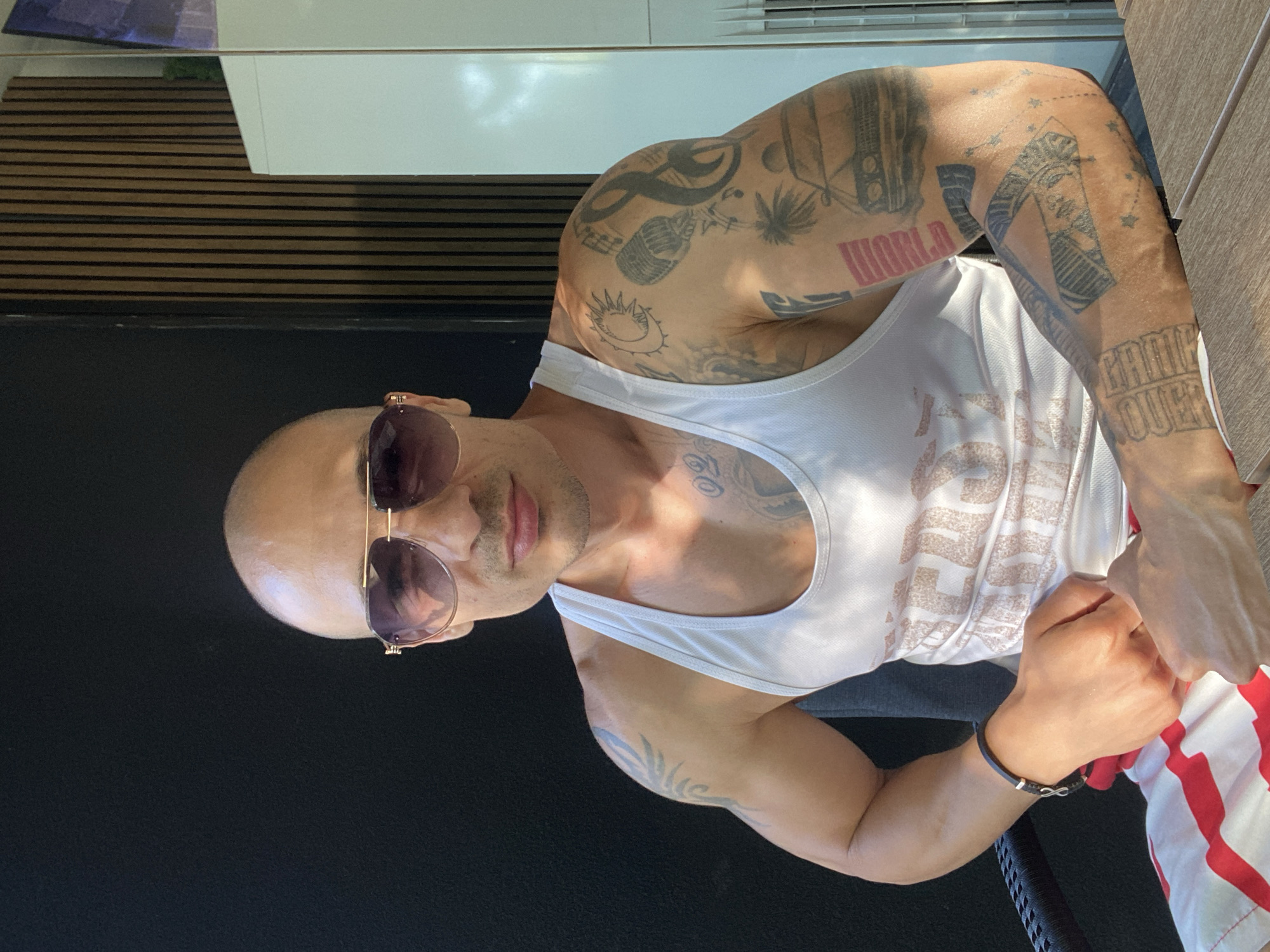 A medium shot captures a bald man, appearing to be of Mediterranean or Middle Eastern descent, seated outdoors. He is wearing a white tank top and sunglasses with gold aviator frames. The man has numerous tattoos on his arms, including depictions of a car, musical notes, a sun, and various abstract designs. His left arm is adorned with a black bracelet featuring an infinity symbol. The background consists of a black wall to the left, and a wall with vertical wooden slats and a white cabinet to the right. Sunlight casts shadows across the scene, suggesting it is daytime. The man is positioned in a chair, with his hands clasped in his lap. His lower body is partially visible, wearing red and white striped shorts. The setting appears to be a modern outdoor patio or balcony. There is no visible indication of the location being in Pleven, Bulgaria.