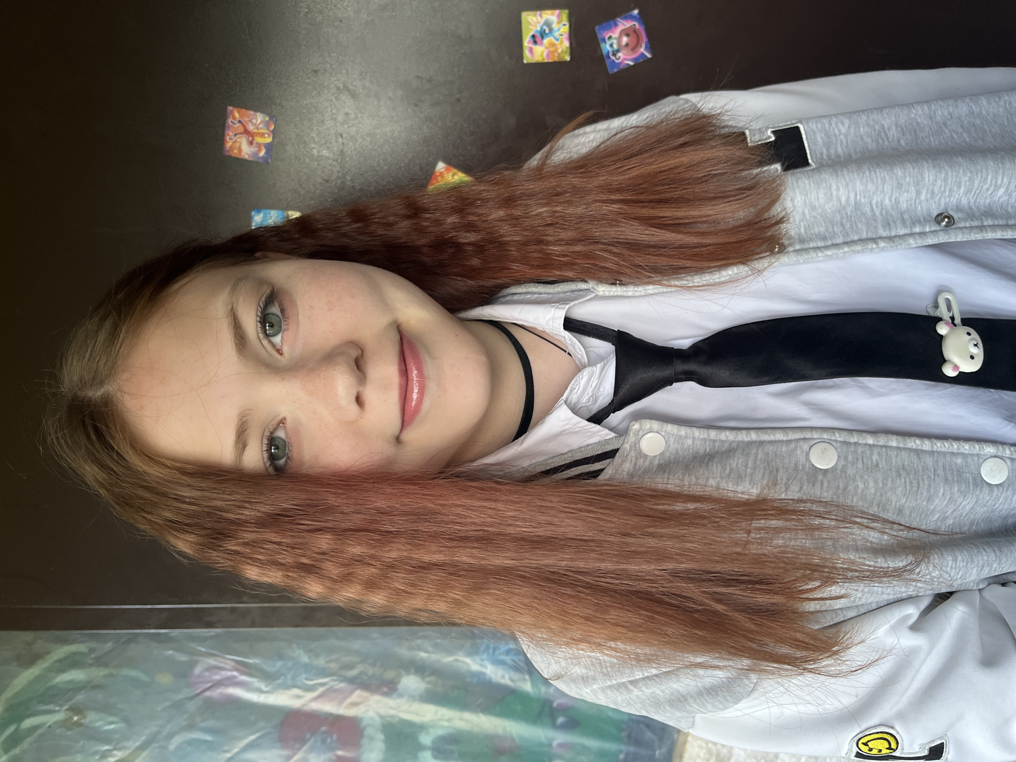 The image captures a young woman in a rotated 90-degree clockwise orientation, presenting her head on the left side of the frame and her body extending downwards. She looks directly towards the right, offering a soft, friendly smile. Her long, reddish-brown hair has a distinct crimped or wavy texture, parted down the middle and falling over her shoulders. She possesses light-colored eyes, appearing blue or green, and her lips are glossy pink.
She is dressed in a white collared shirt, accessorized with a loose black tie and a thin black choker necklace. Over her shirt, she wears a gray and white letterman-style jacket, fastened with snap buttons. A small, white, rabbit-shaped charm, featuring black eyes and a mouth, is clipped onto her black tie. On the lower right part of her jacket, a yellow patch with a black smiley face symbol is clearly visible. Near her head, a partially visible black patch or letter (possibly 'N' or a similar blocky symbol) is seen on the jacket.
The background behind her head and upper body is a dark, plain surface, likely a wall, adorned with several small, colorful square stickers. One sticker above her head depicts an orange and red, possibly creature-like figure, while another to her right shows a blue cartoon character. The very bottom of the image reveals a blurry, colorful surface, possibly a patterned tablecloth or mat. The lighting appears natural, suggesting daytime. The overall impression is a casual and relaxed portrait taken in an indoor setting. No discernible text is visible beyond the symbolic facial features on the charm and patch.