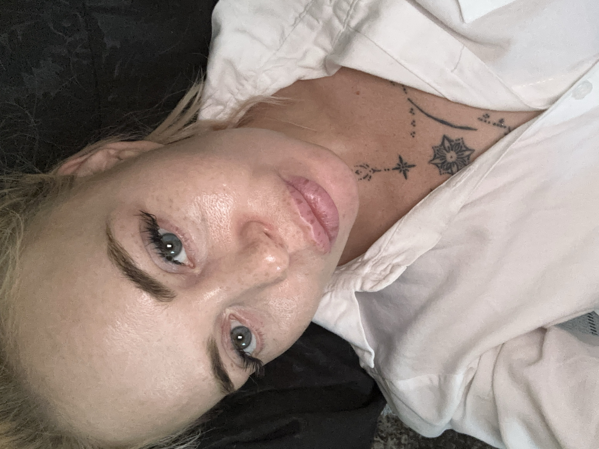 A close-up, high-angle shot shows a blonde woman in a white collared shirt. She has light blue eyes, full lips, and visible freckles on her nose and cheeks. False eyelashes are applied. She has a tattoo on her chest with a starburst design and other small dots and lines. The background is dark and indistinct. The lighting appears to be from above and to the side.