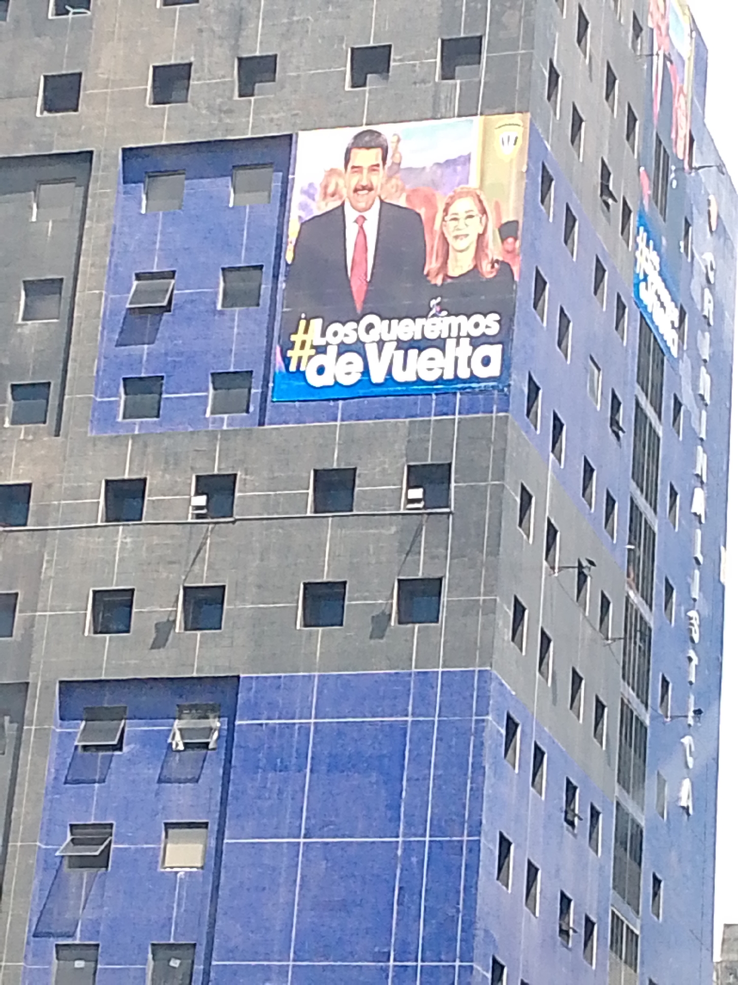 This image captures a tall building in Caracas, Venezuela, under clear daylight conditions. The building features a prominent large billboard displaying two smiling individuals: a man, identifiable as Nicolás Maduro, and a woman, identifiable as Cilia Flores. Maduro is depicted wearing a suit and red tie, while Flores wears a black top.
Beneath their portraits, large white text with a blue background reads "¡Los Queremos #de Vuelta" which translates to "We want them #back." This is a political slogan, likely part of a campaign.
The building itself is multi-storied and appears to be an office or residential block. Its facade is a mosaic of grey and blue panels, with the blue sections appearing on the lower parts of the visible side and also vertically on one corner. Numerous square windows are visible, many of which are open, some with their awnings or top-hinged sections extended outwards, creating shadows on the building's surface. The bright sunlight suggests a sunny day. The overall impression is a vibrant political message integrated into the urban landscape.