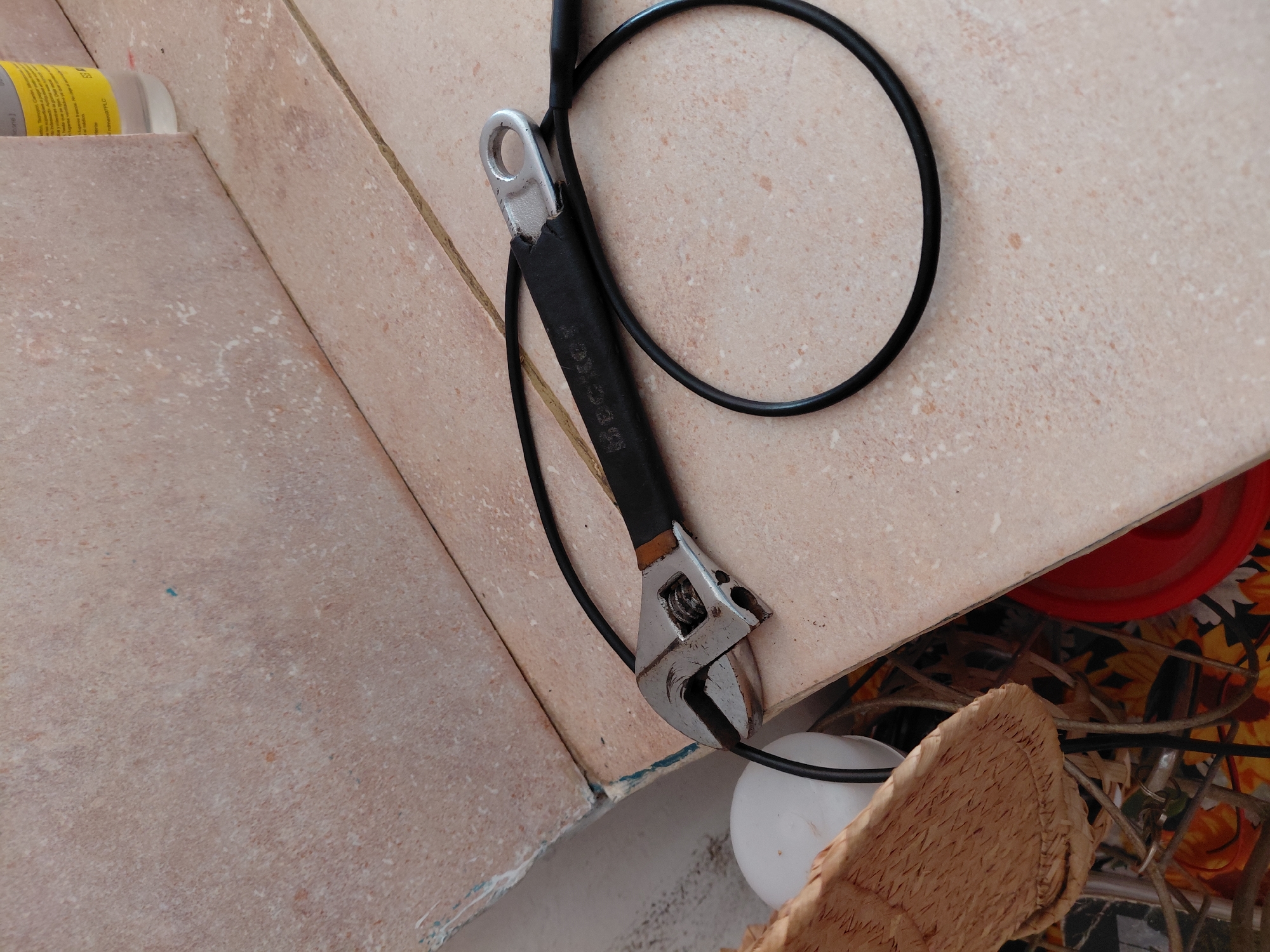 This close-up image displays a collection of tools and miscellaneous items resting on and around a light-colored, speckled tile surface. The setting appears to be an indoor or semi-sheltered utility area, possibly a workbench or storage shelf, in Santa Ana, Venezuela.
The central object is a black-handled adjustable wrench (monkey wrench), positioned diagonally across the main tiled surface. Its silver-colored metal head, including the adjustable jaw mechanism, is prominent. The black handle bears some faint, embossed text, partially visible as "SEC-ROY" or similar. A black electrical cable or hose is loosely coiled around the wrench and extends across the tile surface, forming a large loop.
To the left of the wrench, a section of a yellow bottle with small, indistinct white text is visible, resting on an adjoining tiled surface. Below the edge of the main tiled surface, a cluttered area is revealed. Here, a white spherical object, a piece of light-brown woven material (resembling a mat or basket), a bright red plastic object, and a floral pattern fabric (featuring yellow sunflowers) can be seen. The lighting in the image suggests it was taken during daylight hours. There is no discernible activity taking place; the objects appear to be static, possibly stored or temporarily left in place. The tiles show some signs of wear and minor discoloration.