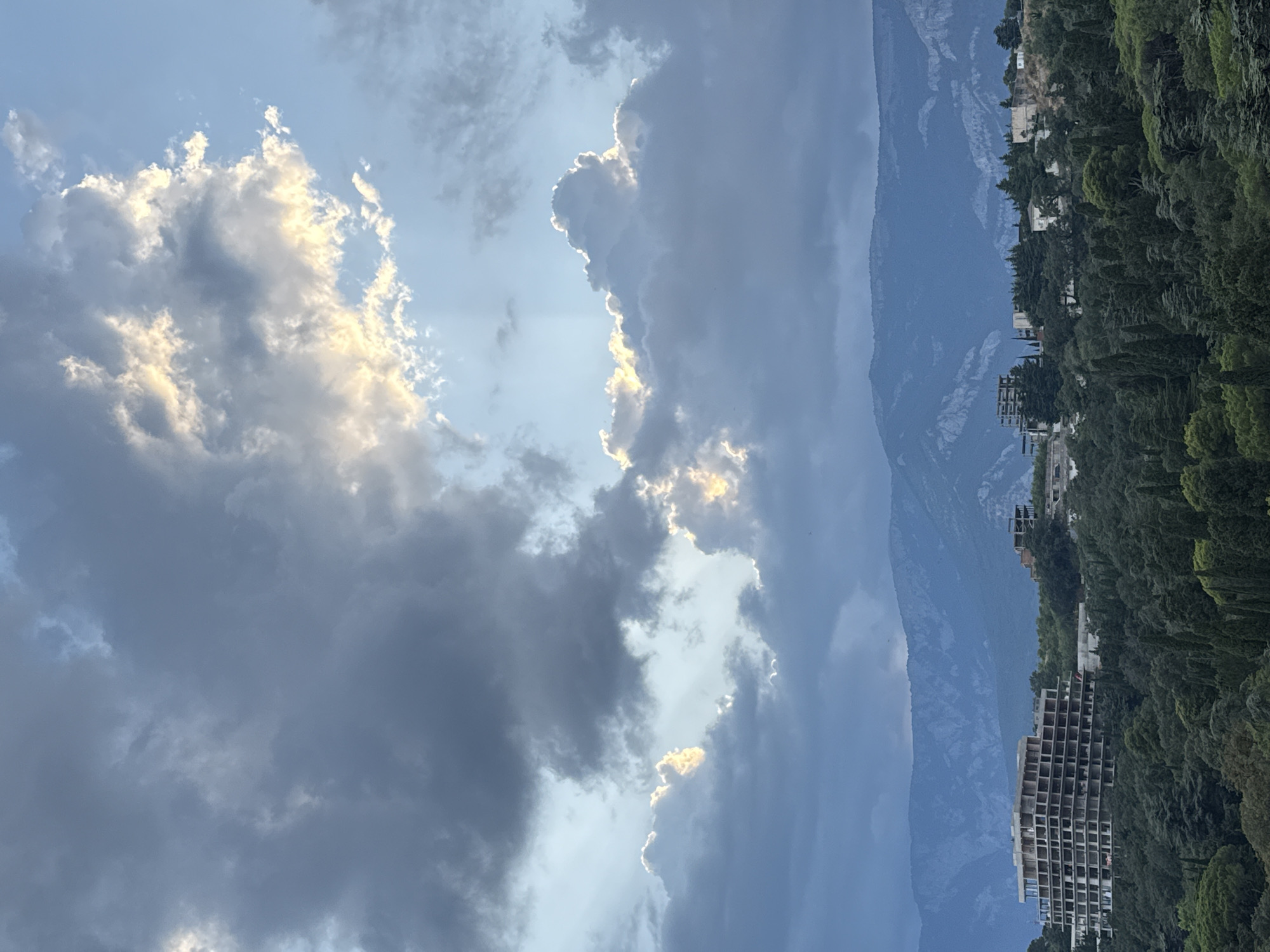 The image captures a scenic view overlooking the city of Yalta, Ukraine, with a focus on the dramatic interplay of light and shadow in the sky above a lush, green landscape. In the foreground, a dense forest of various trees, including tall cypress trees, covers the rolling hills. Scattered amongst the greenery are several unfinished or abandoned buildings, one of which is a large, multi-story concrete structure with exposed framework. In the distance, a range of majestic mountains forms a stark silhouette against the sky, their peaks appearing weathered and rugged. The sky is a canvas of dynamic clouds, with patches of bright sunlight breaking through the darker, ominous storm clouds, creating a sense of both drama and beauty. The overall impression is one of nature's grandeur contrasted with the presence of human development, some of which appears to be in a state of disrepair.