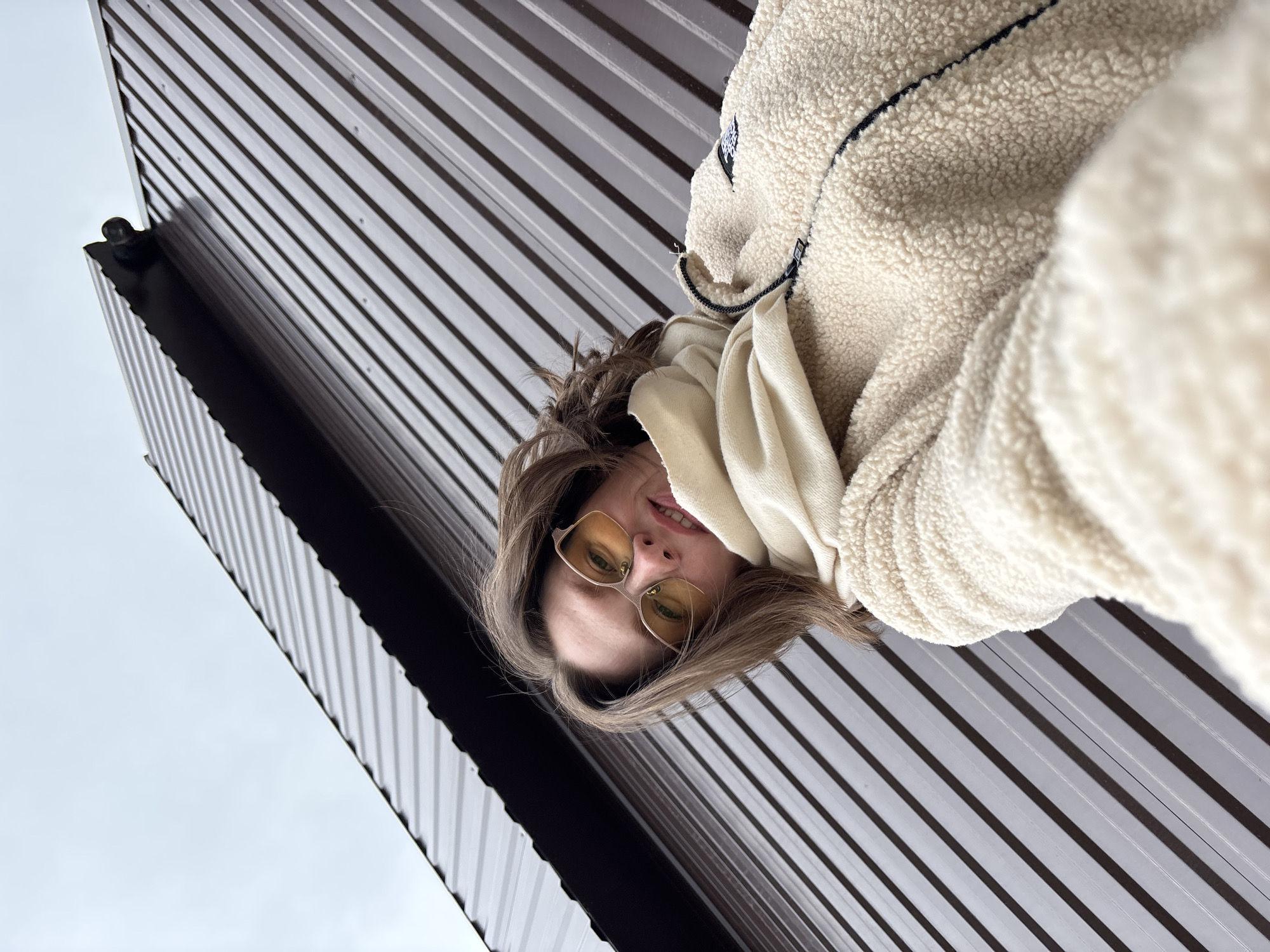 A low-angle selfie depicts a fair-skinned woman with light brown hair and yellow-tinted eyeglasses, wearing a cream-colored fleece jacket and a white scarf. The background consists of a metal roof with a dark brown and gray striped pattern, and a glimpse of a light gray sky. The woman appears to be looking down at the camera. The location is Chaikovsky, Russia, evidenced by metadata, if available.