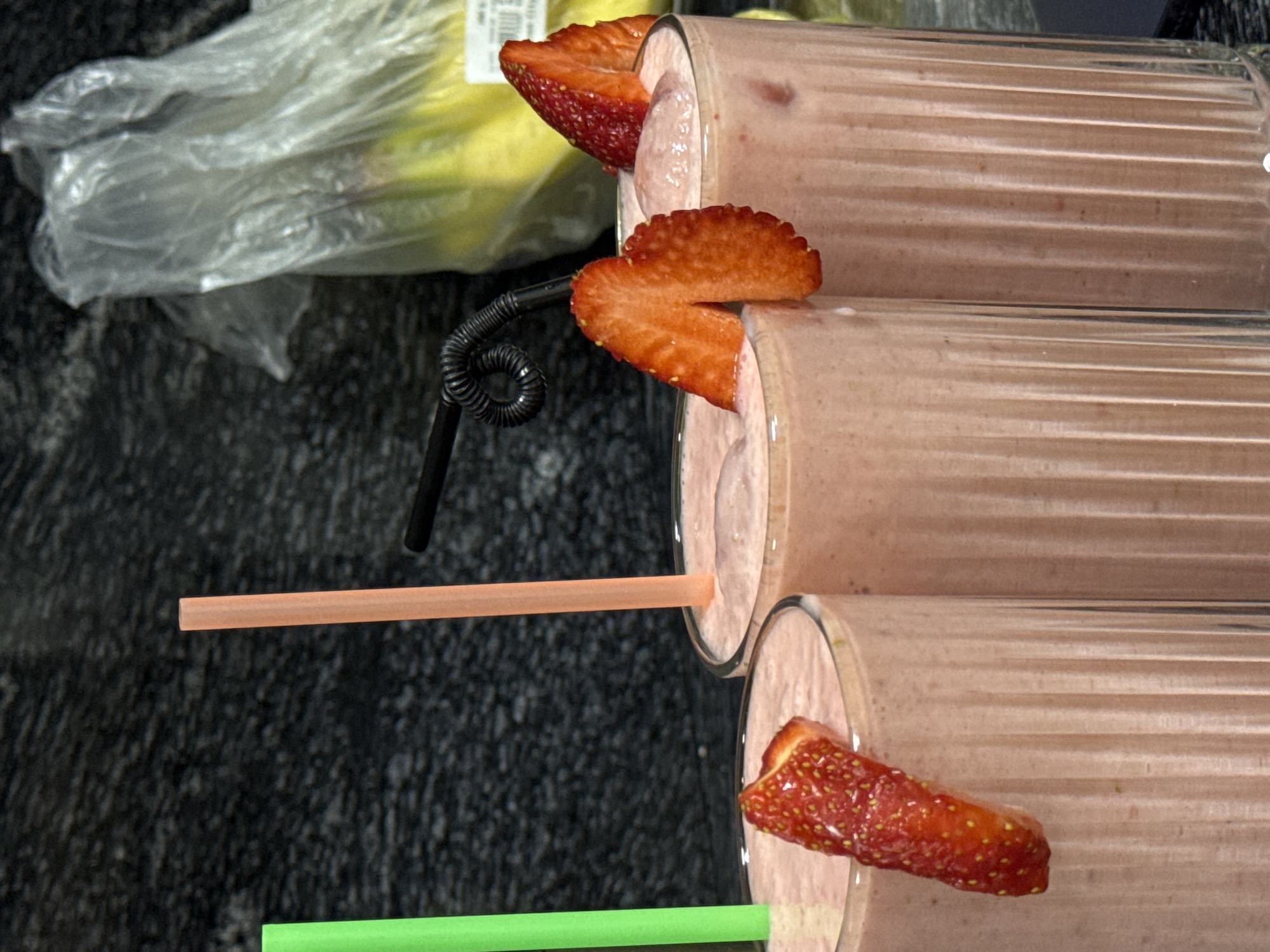 The image depicts three glasses of strawberry smoothies, each garnished with a sliced strawberry and a straw. The straws are different colors: green, orange, and black. Behind the smoothies, partially visible, is a clear plastic bag containing bananas. The background is a dark, speckled surface. The location and specific details suggesting it is in Yerevan, Armenia, are not evident in the image.