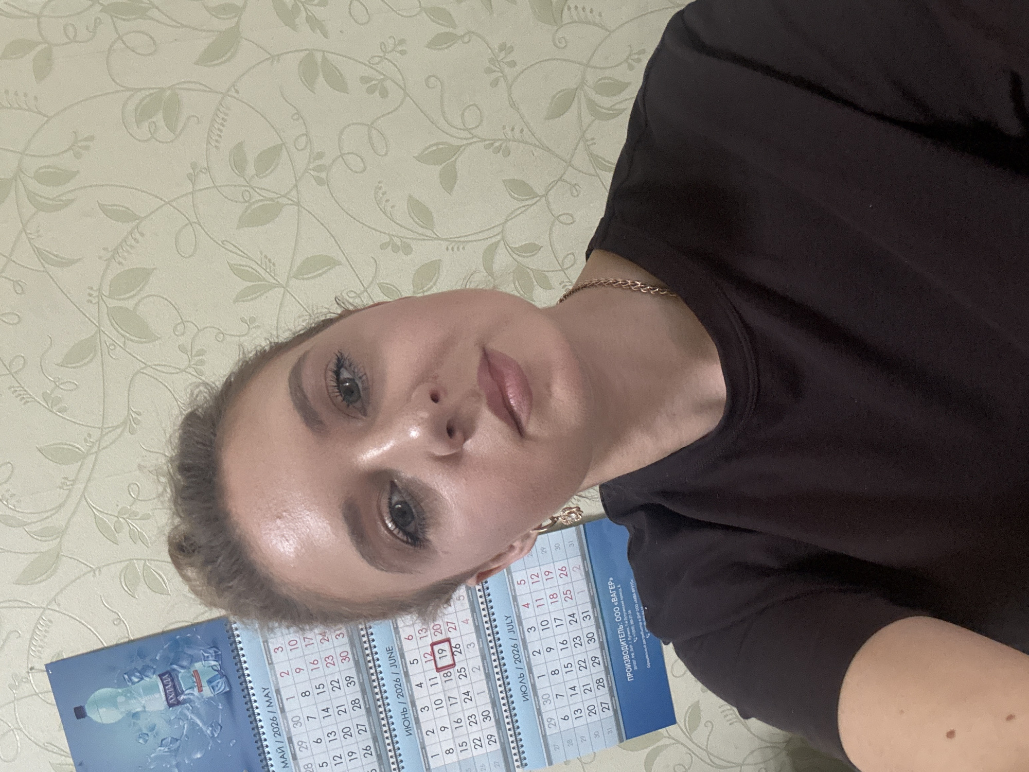 A woman with blonde hair pulled into a bun is shown from the chest up, taking a selfie. She is wearing a dark t-shirt and a gold chain necklace. Her gaze is directed at the camera. In the background, a floral wallpaper is visible, and to the left, a calendar is hanging on the wall, displaying dates for May, June, and July of 2026.