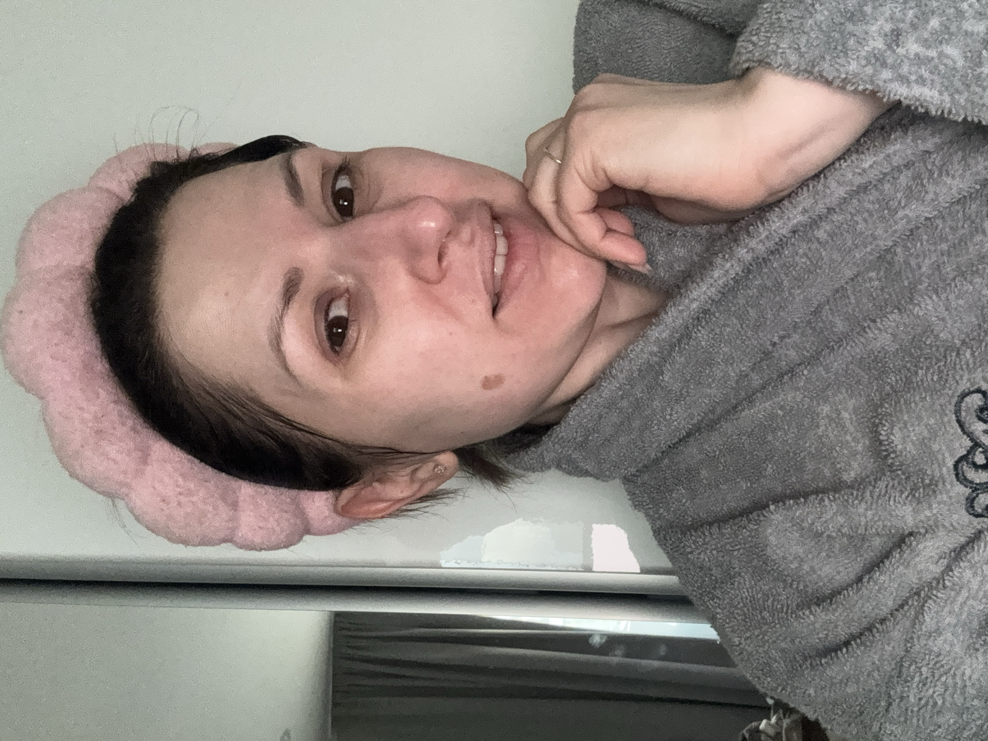 A woman is pictured indoors, likely in a domestic setting. She is wearing a pink headband and a gray, textured robe. Her left hand is raised to her face, with a ring visible on one finger. Her skin tone is fair, and a mole is present on her cheek. The background appears to be a white, possibly reflective surface, suggesting a refrigerator or similar appliance. The image does not provide sufficient details to determine a location within Sofia, Bulgaria.