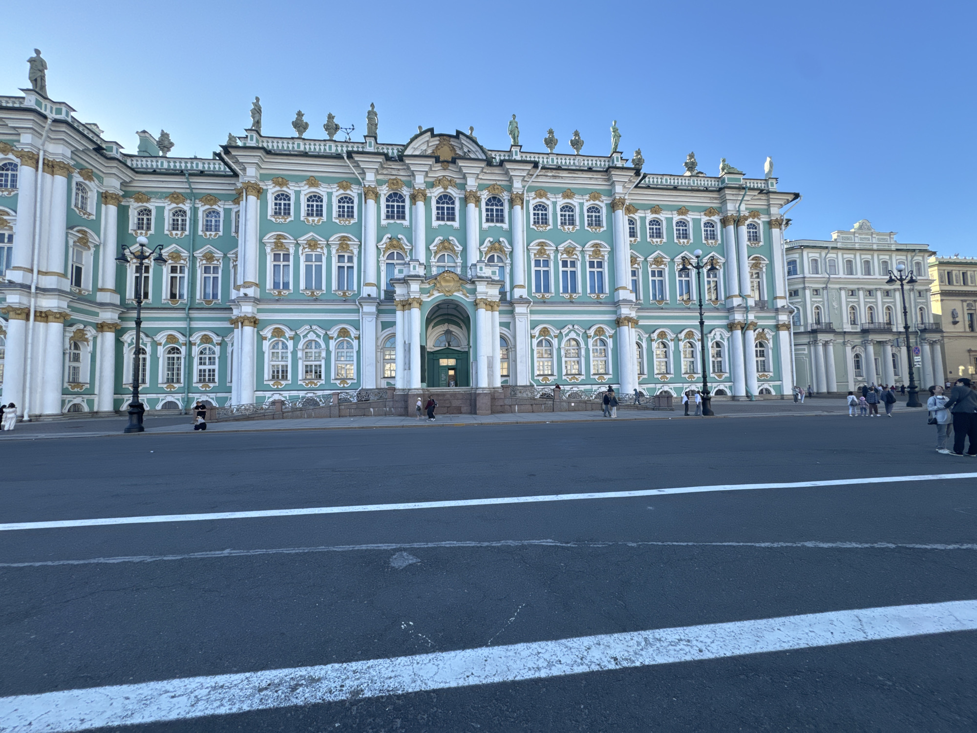 The image depicts a wide, bright view of the State Hermitage Museum, formerly the Winter Palace, in Saint Petersburg, Russia. The iconic turquoise and white facade of the palace dominates the center of the frame, adorned with gold accents and numerous windows. Statues are visible atop the building's roofline and along its facade. In front of the palace, a large paved area, likely Palace Square, is visible, with white lines marking a crosswalk in the foreground. Several people are scattered across the paved area, some walking and others standing in groups. A few lampposts are also present. The sky is a clear, vibrant blue, indicating a sunny day. The overall scene is one of historical grandeur and public space.