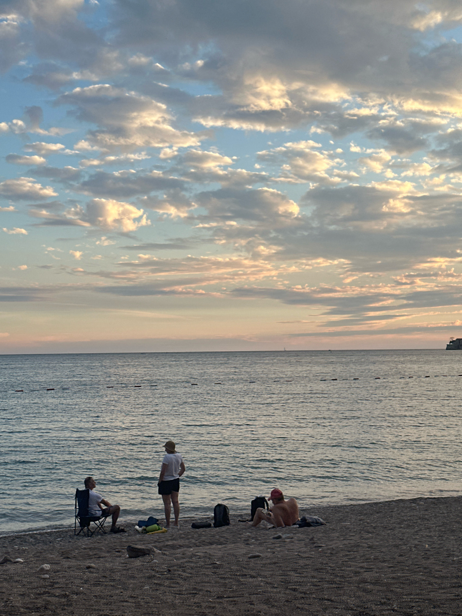 The image captures a serene beach scene in Budva, Montenegro, during what appears to be either sunrise or sunset, indicated by the soft, warm hues in the sky and the gentle undulation of the sea. The sky is adorned with scattered clouds, painted in shades of white, gray, and delicate pinks, contrasting with the tranquil blue of the upper atmosphere.
On the pebbled beach, three individuals are visible. To the left, a person is seated in a dark-colored folding chair, facing the water, wearing a white t-shirt and shorts. Adjacent to them, another person stands, also in a white t-shirt and dark shorts, looking towards the sea. A straw hat is perched on their head. To the right, a third person is seated on the sand, wearing a red cap and appearing to be bare-chested. Various items, including backpacks and clothing items, are scattered on the beach near the individuals.
The sea stretches out to the horizon, with a calm surface reflecting the sky's colors. A line of buoys or markers is visible across the water, suggesting a designated swimming area or a barrier. In the distance, on the far right of the horizon, a structure that appears to be a building or a fortification is perched on a cliff, hinting at the historical context of Budva. The overall atmosphere is one of peaceful relaxation by the Adriatic Sea.