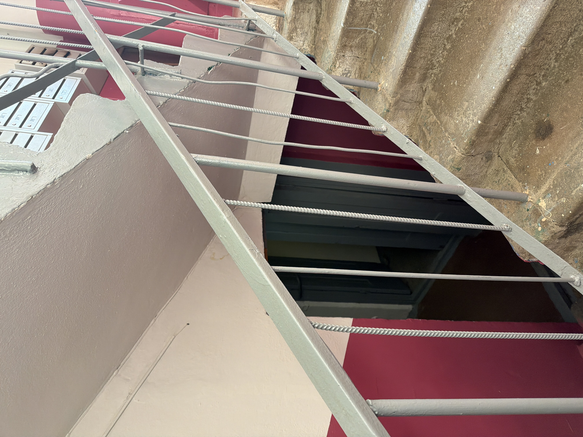 This is an eye-level, medium shot of a concrete staircase in a Soviet-era apartment building in Okha, Russia. The staircase is weathered and stained, with a metal railing that is also worn. The walls are painted a light pink and a dark red. In the background, there is an open doorway leading to a dark interior. There are also several mailboxes visible at the top of the stairs. The lighting is dim, suggesting it is either a cloudy day or the staircase is located indoors.