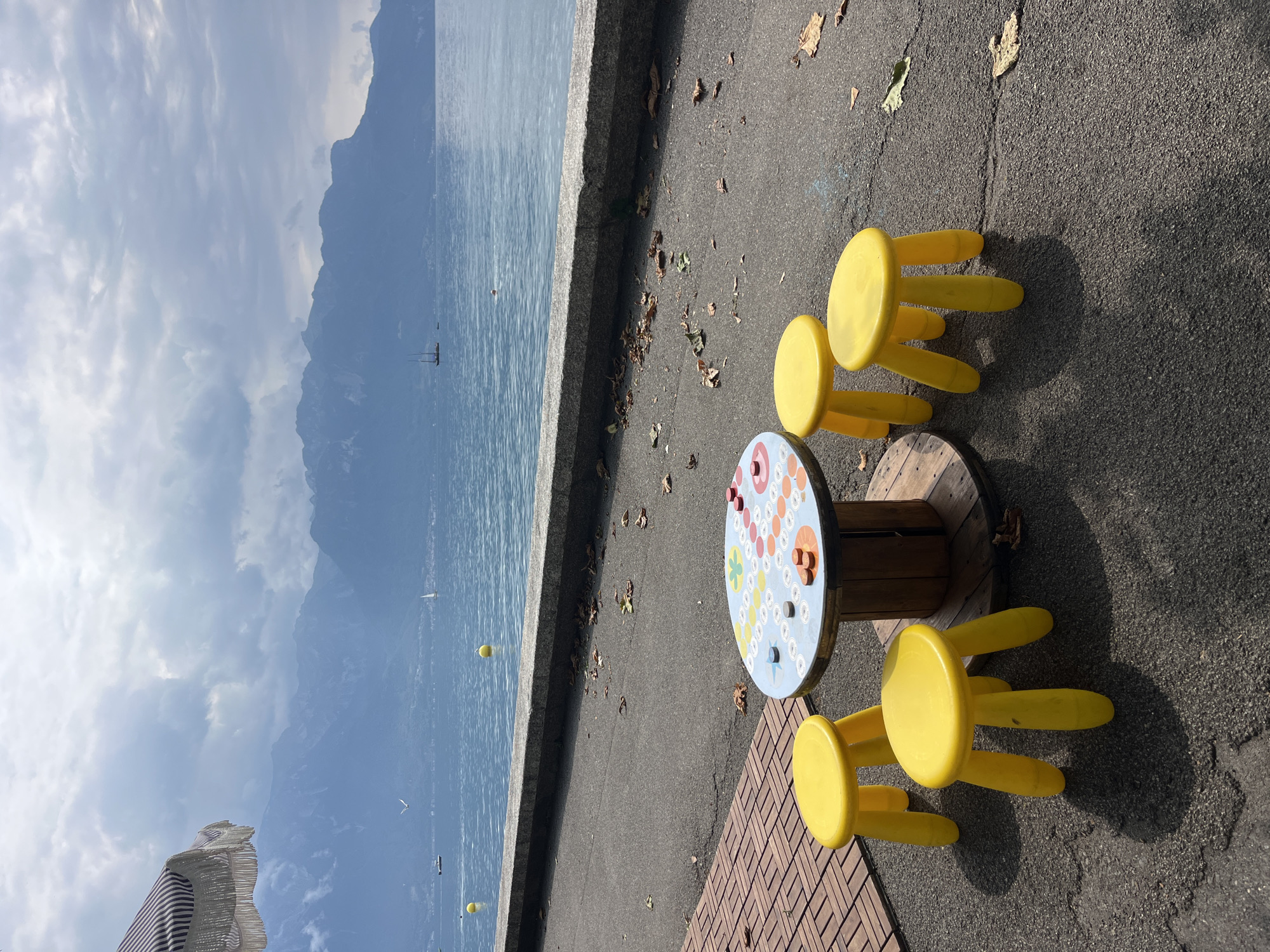 The image depicts a scene near the shore of Lake Geneva in La Tour-de-Peilz, Switzerland. In the foreground, a circular table with a board game printed on its surface stands on a wooden spool. Five bright yellow stools are arranged around the table, suggesting a recreational area for children or a game space. The ground is paved with asphalt, and scattered autumn leaves are visible. To the left, a portion of a striped umbrella is visible, offering shade.
In the middle ground, a concrete walkway or embankment separates the paved area from the lake. The lake itself is calm and blue, with a few yellow buoys floating on the surface. In the distance, sailboats can be seen on the water.
The background is dominated by a range of majestic mountains, their peaks partly obscured by dramatic, cloudy skies. The overall atmosphere suggests a slightly overcast day, possibly in the autumn. The view is characteristic of the landscape found along the shores of Lake Geneva, with the Alps providing a stunning backdrop.