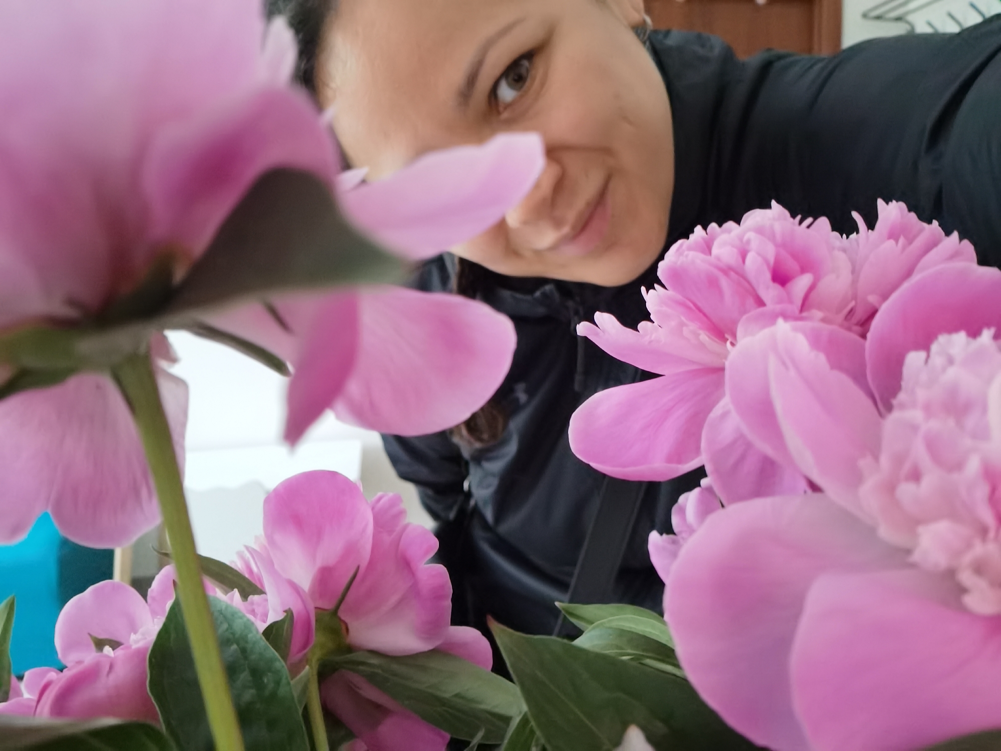 The image presents a close-up, intimate portrait of a woman peeking from behind a vibrant display of pink peonies. The main subject is an adult woman with dark hair, visible on the right side of the frame. Her face is partially shown, tilted slightly, with a gentle, soft smile and warm brown eyes looking directly towards the viewer. She is wearing a dark, possibly black, jacket or top.
The foreground and midground are filled with numerous pink peonies, some in sharp focus revealing their delicate petals and ruffled texture, while others are softly blurred, creating a sense of depth and framing the woman's face. Green leaves are interspersed among the blossoms. The overall composition suggests a casual and possibly playful moment, perhaps a selfie or a close-up portrait taken with the flowers.
The background is softly out of focus, indicating an indoor setting with light-colored walls or surfaces and indistinct elements, possibly furniture, further back. The lighting appears natural and soft, contributing to the pleasant and gentle mood of the scene. No text is visible in the image. The context for the photo is Chaikovsky, Russia, though no specific visual cues within the image itself confirm this location.